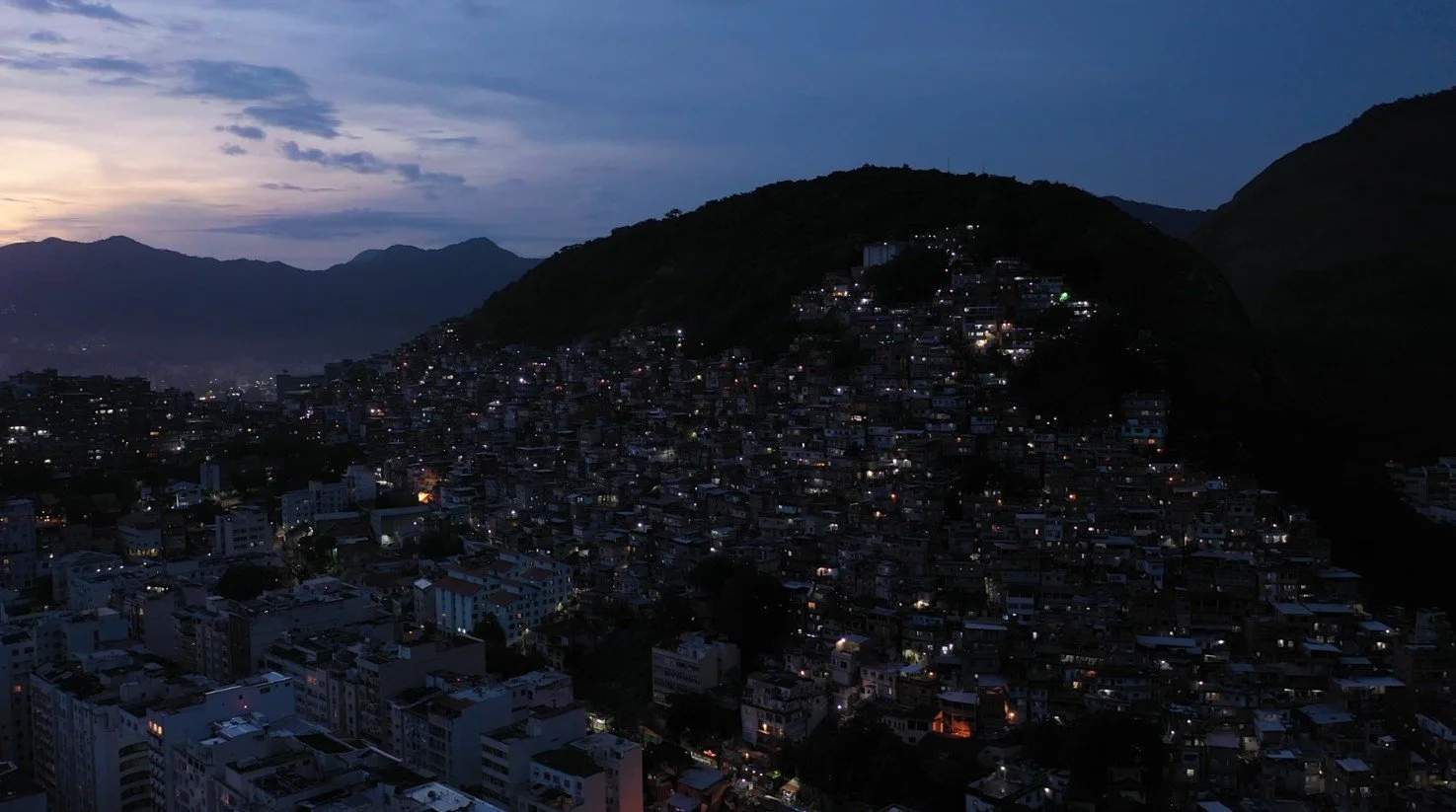 An aerial twilight view of a densely packed hillside neighborhood, with countless small homes lit by scattered lights, surrounded by dark mountain silhouettes under a cloudy evening sky.