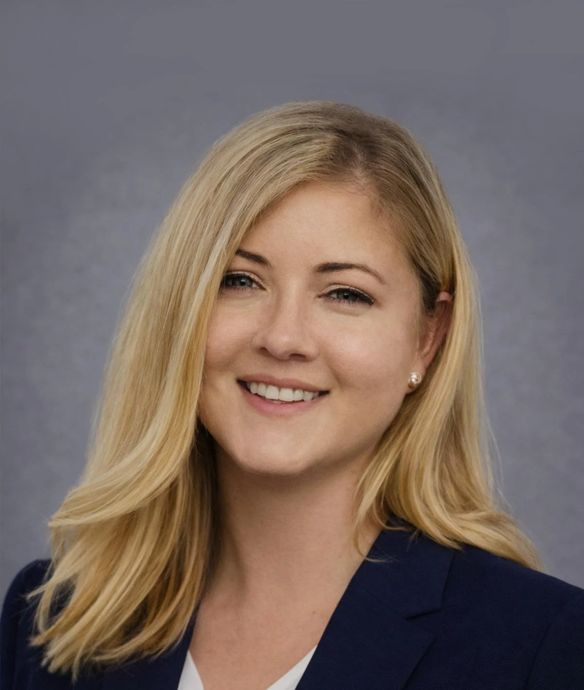 A young woman with blonde hair styled in loose waves, wearing a navy blazer and pearl earrings, smiling against a gray background.
