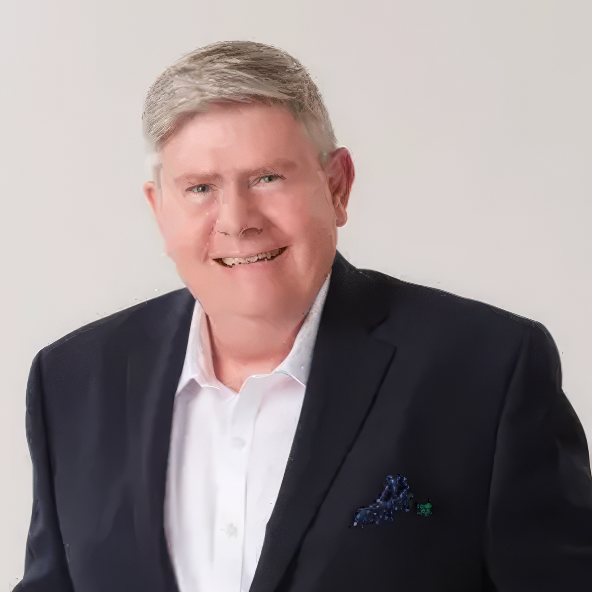 Portrait of a smiling man with gray hair, wearing a black suit jacket, white shirt, and a pocket square, against a plain white background.