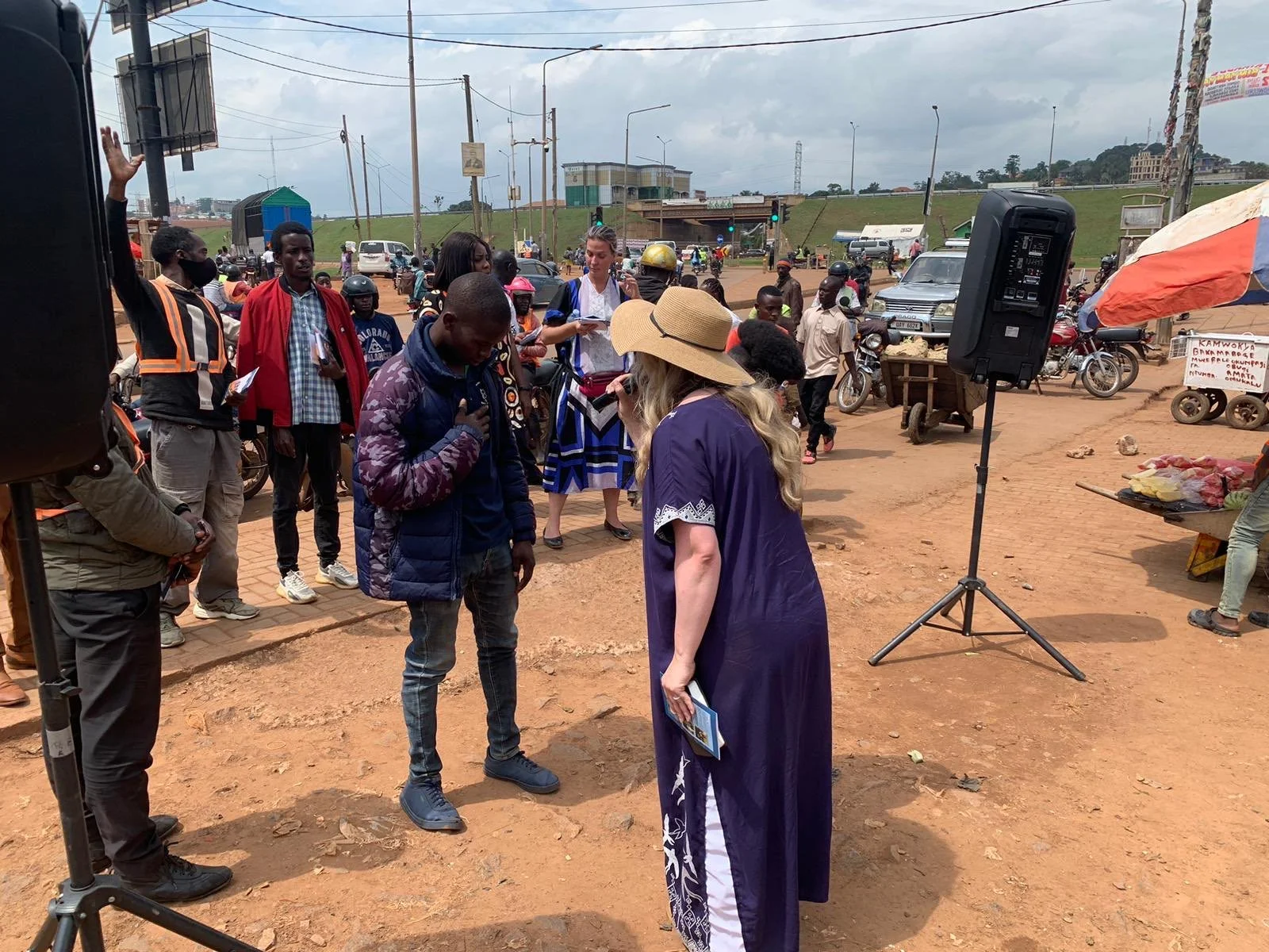 A woman in a wide-brim hat and long blue dress speaks into a microphone to a man standing nearby on a dusty roadside, while people, motorcycles, and vehicles gather around under a cloudy sky with power lines and buildings in the distance.