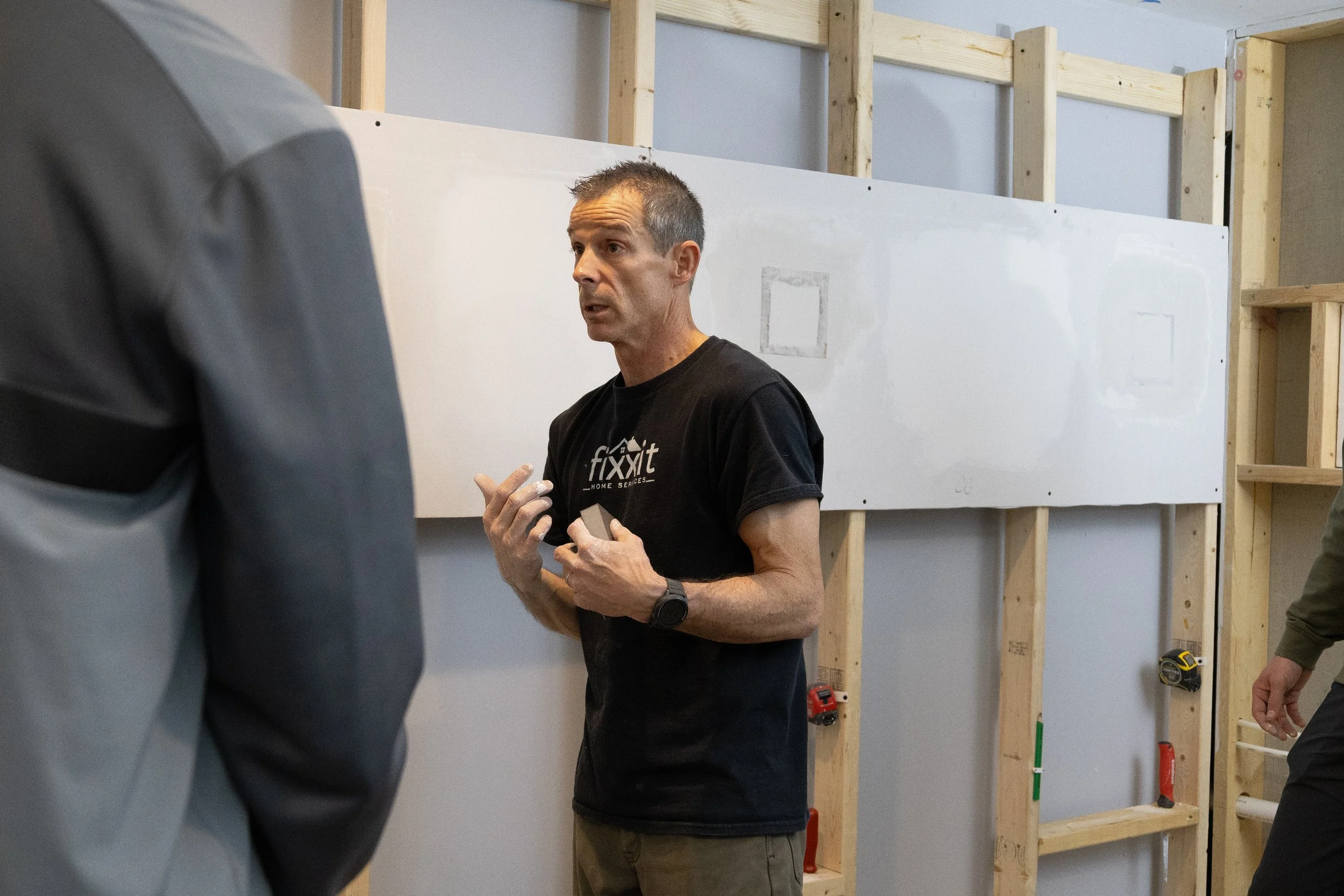 A man in a black t-shirt with 'fixxit' logo is talking to two other people in a construction space with unfinished wall framing and tools.