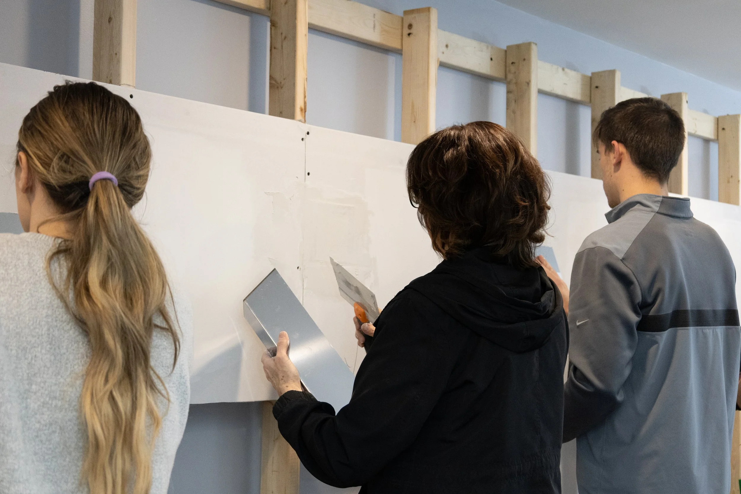 Three people working on drywall installation, with two holding putty knives and one standing nearby, in a room under construction.