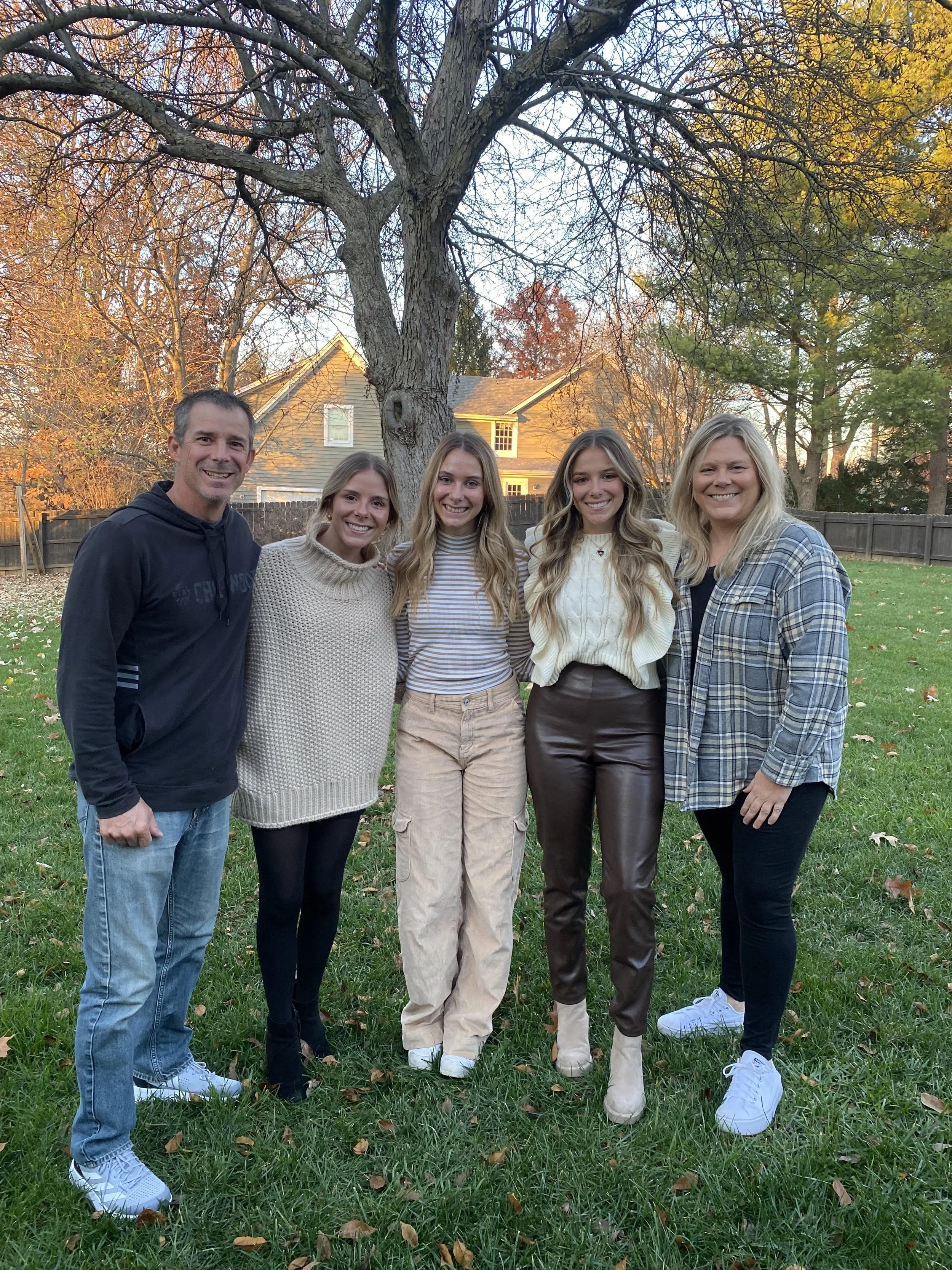 A group of five people, two men and three women, standing outdoors on grass in front of a large tree with fall-colored leaves, smiling for the camera, with houses and trees in the background during late afternoon or early evening.