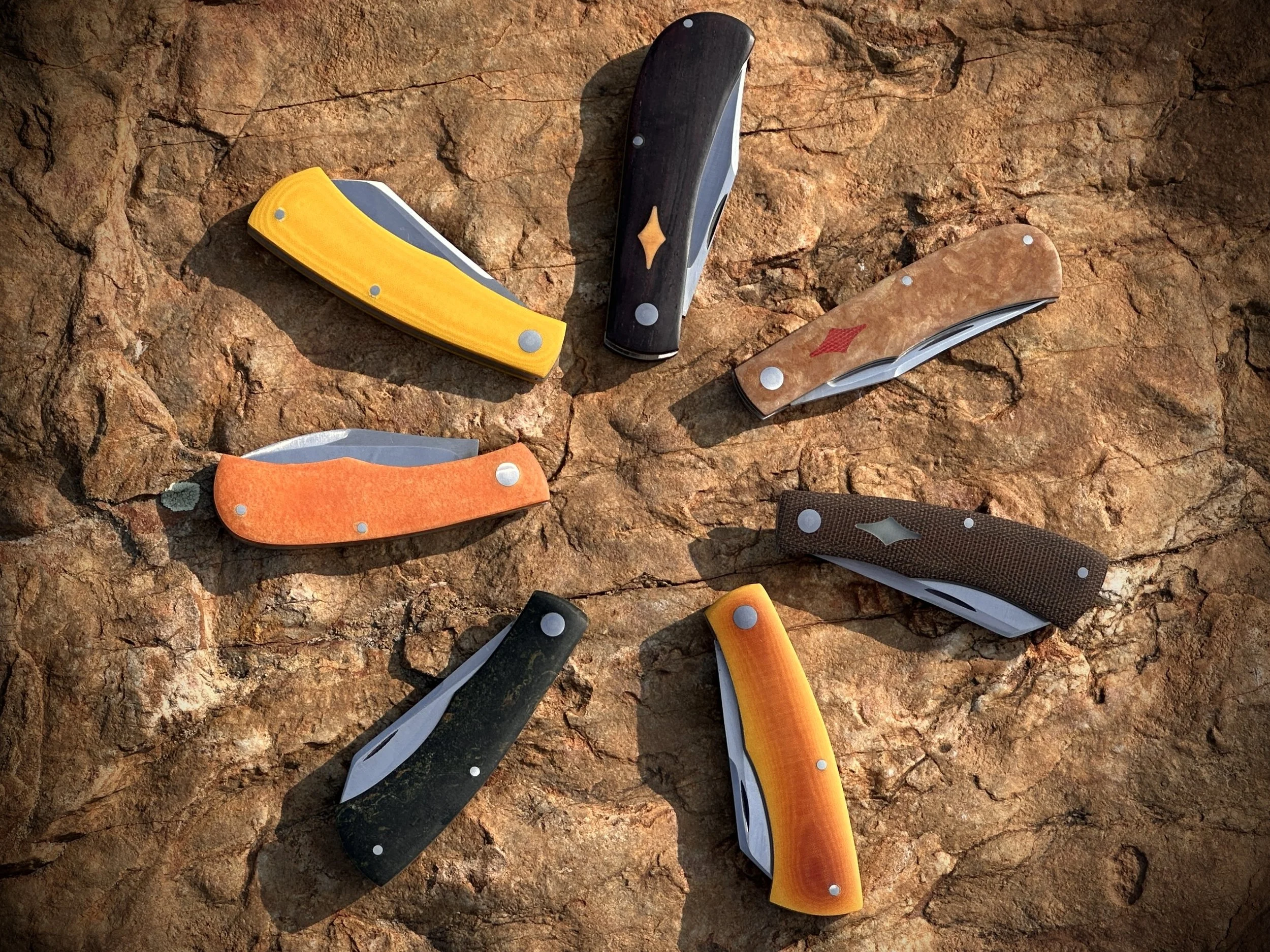 Eight folding pocket knives arranged in a circle on a rocky surface, each with a different colored handle.