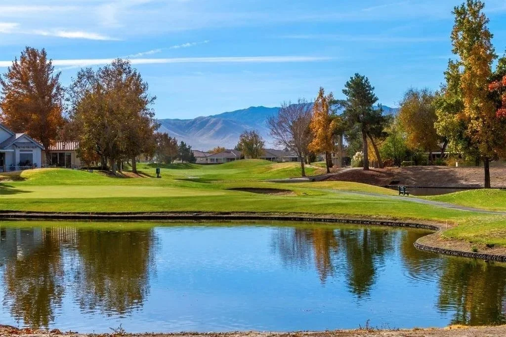 A scenic golf course with green fairways, trees in autumn colors, a small pond reflecting the sky, and mountains in the background under a blue sky.
