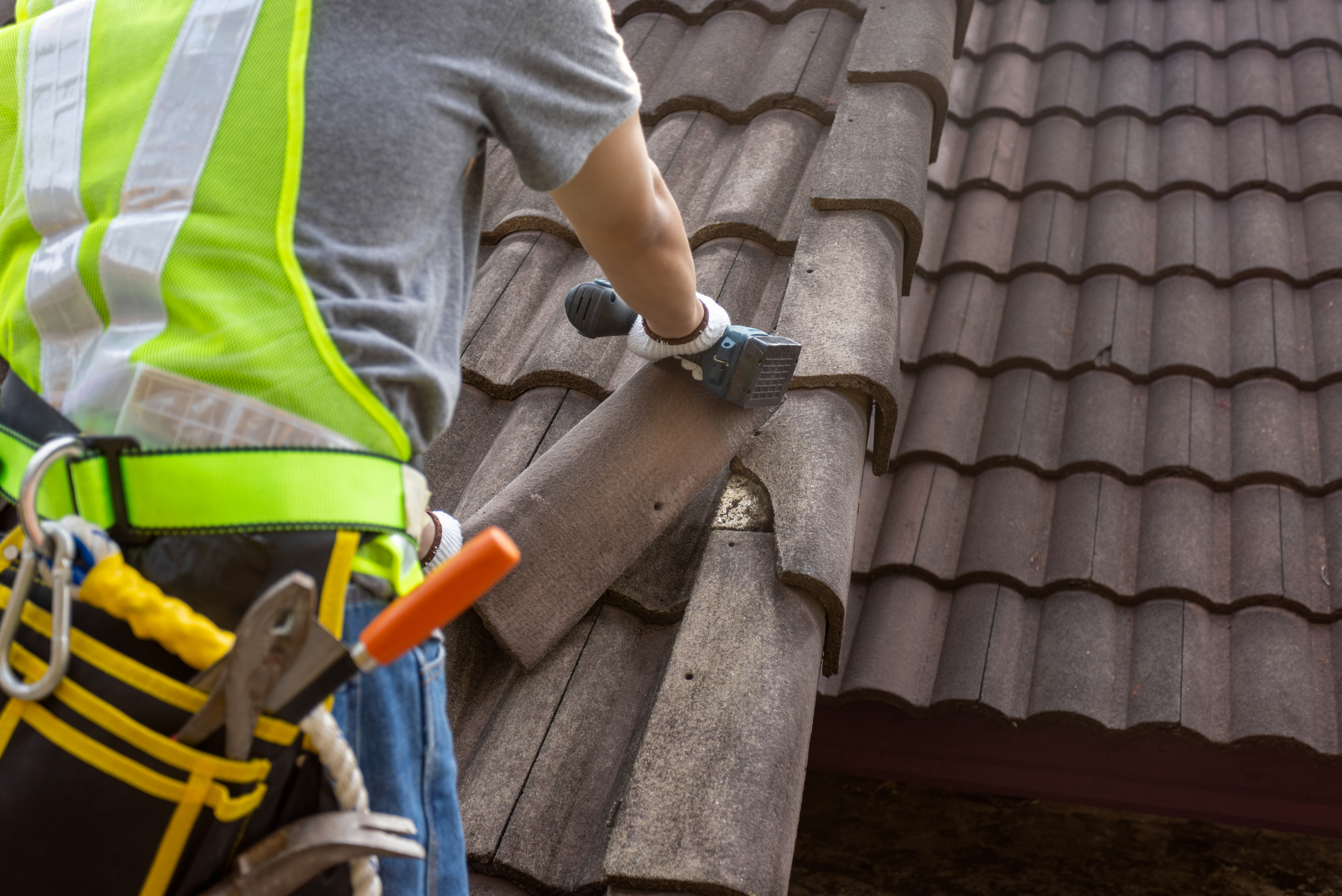 Worker on roof installing or repairing roof tiles, wearing safety gear and holding a cordless drill.