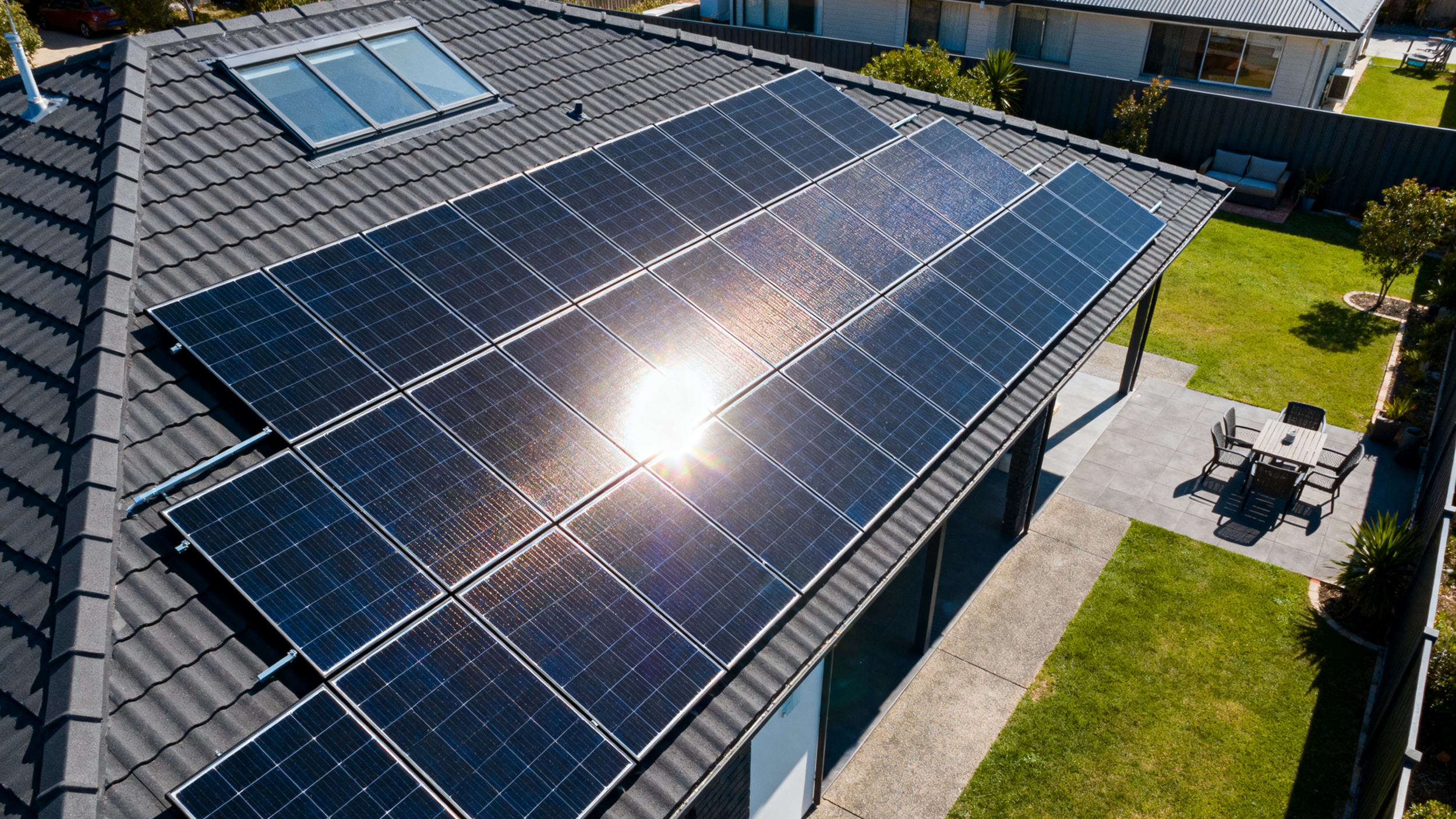 A residential house roof with multiple solar panels installed, reflecting sunlight, in a backyard with a patio table and chairs, grass, and adjacent houses visible.