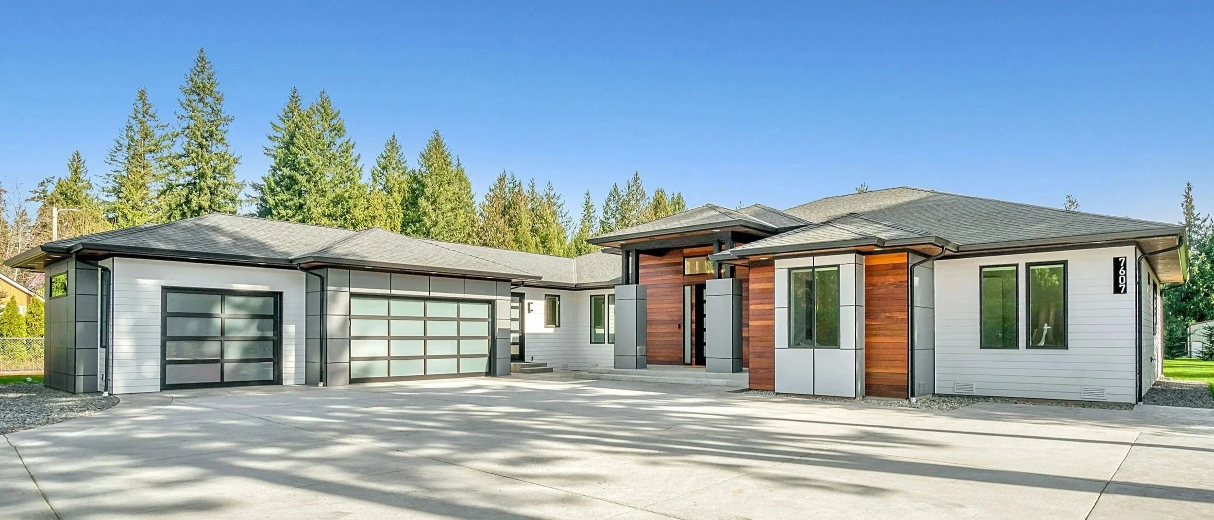 Modern single-story house with white and wood exterior, glass garage doors, concrete driveway, and surrounded by tall trees under a clear blue sky.