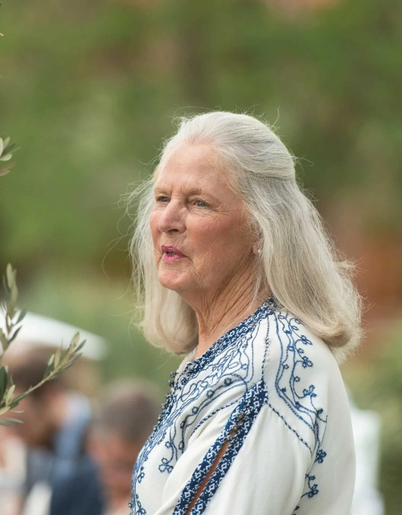 Older woman with long gray hair wearing a cream-colored blouse with blue embroidery, outdoors with blurred greenery and other people in the background.
