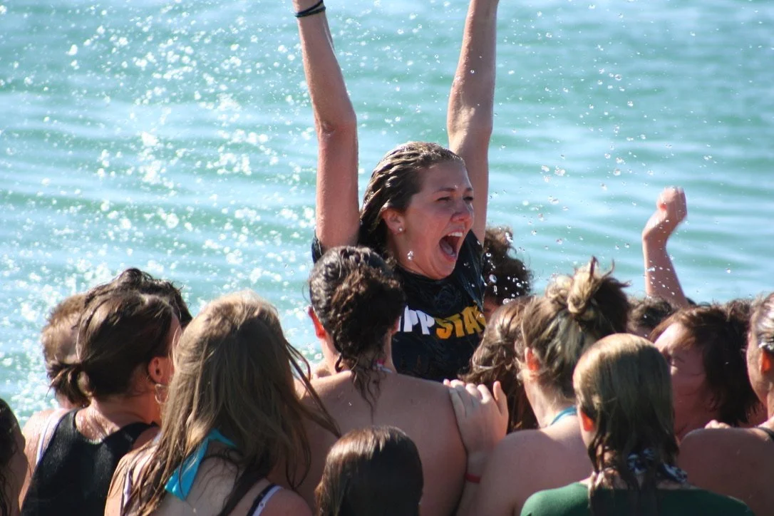 A group of young women celebrating in a body of water, with one woman being lifted above the others, expressing joy and excitement.