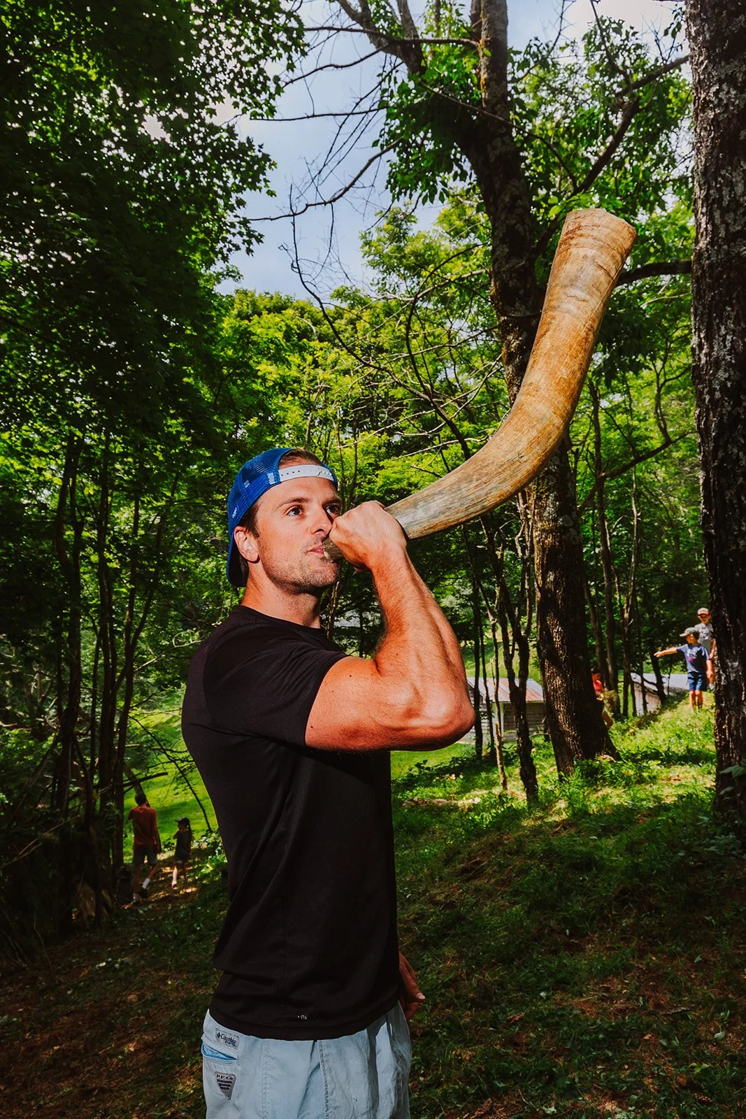 A man in a black shirt and blue cap holding a large horn, appearing to blow into it, in a wooded outdoor area with green foliage and other people in the background.