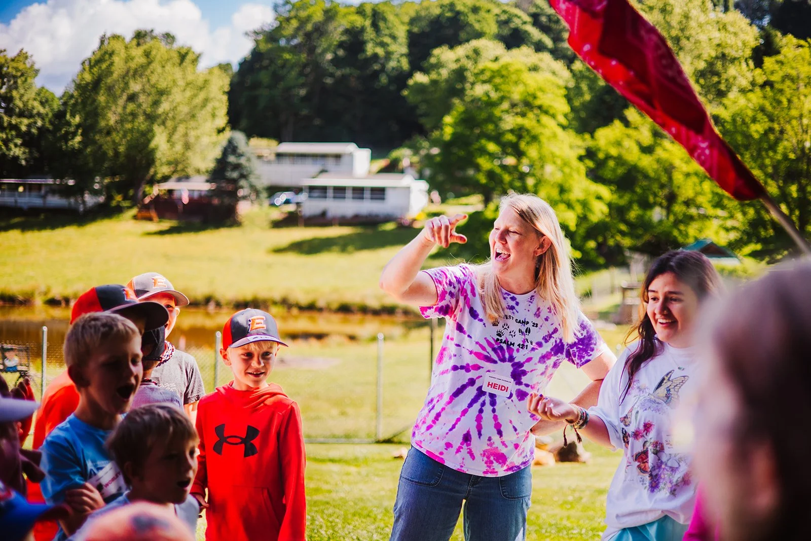 A group of children and a woman are outdoors on a sunny day, participating in an activity or game. The woman is gesturing or giving instructions, and everyone appears engaged and smiling. The setting includes green trees, grass, and a lake or pond in the background.