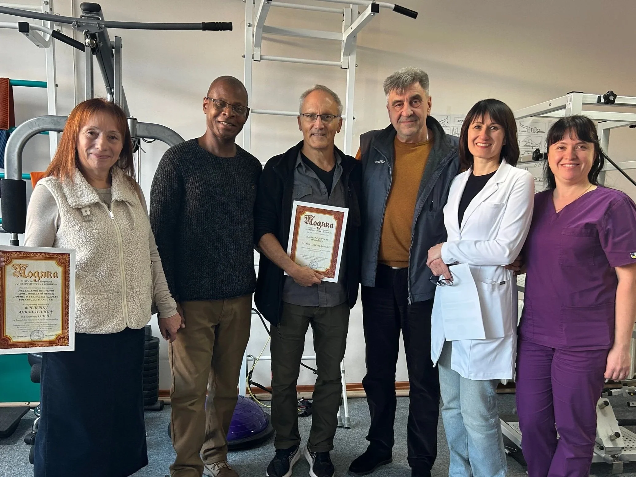 Group of seven people posing together in a gym or rehabilitation center, some holding framed certificates, with exercise equipment in the background.