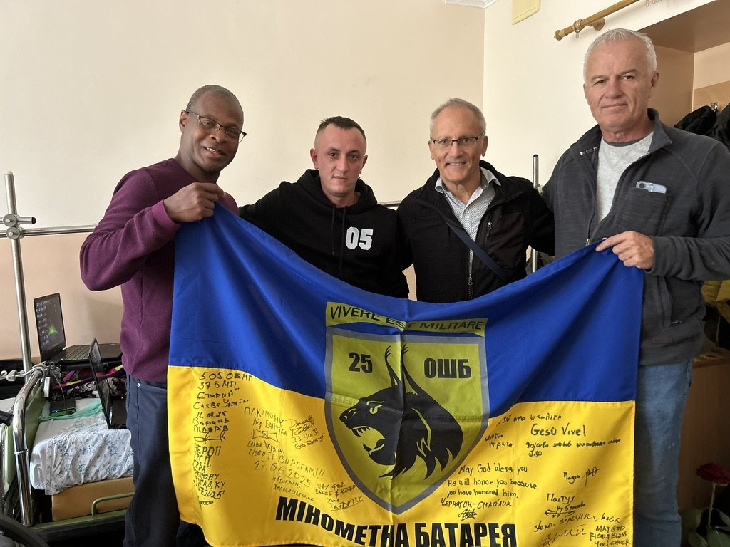 Four men standing indoors, holding a blue and yellow flag with signatures and a panther emblem, celebrating or commemorating a military unit.
