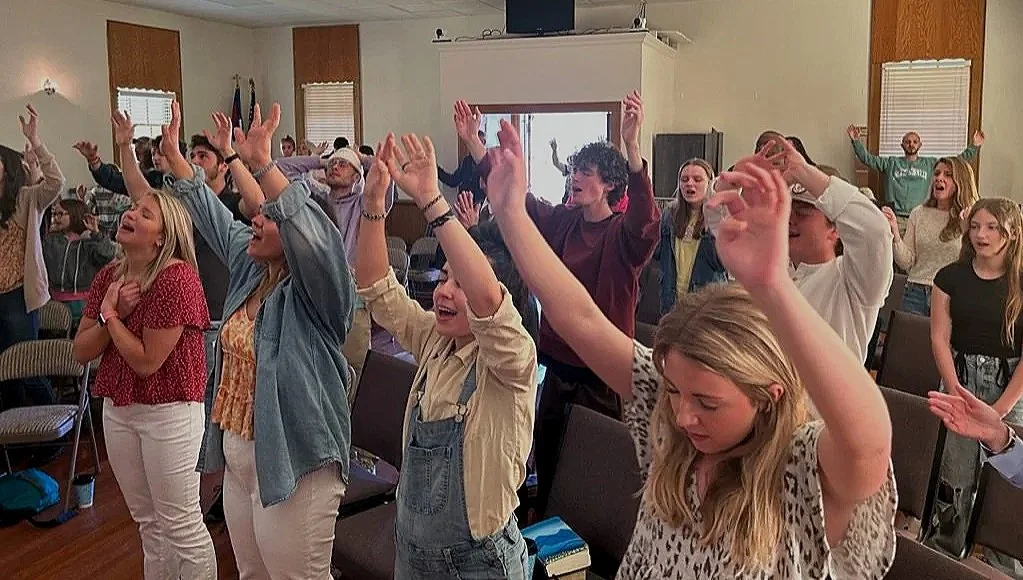 Group of people standing with their hands raised, participating in a joyful activity in a room with beige walls and windows, some smiling and others with eyes closed.