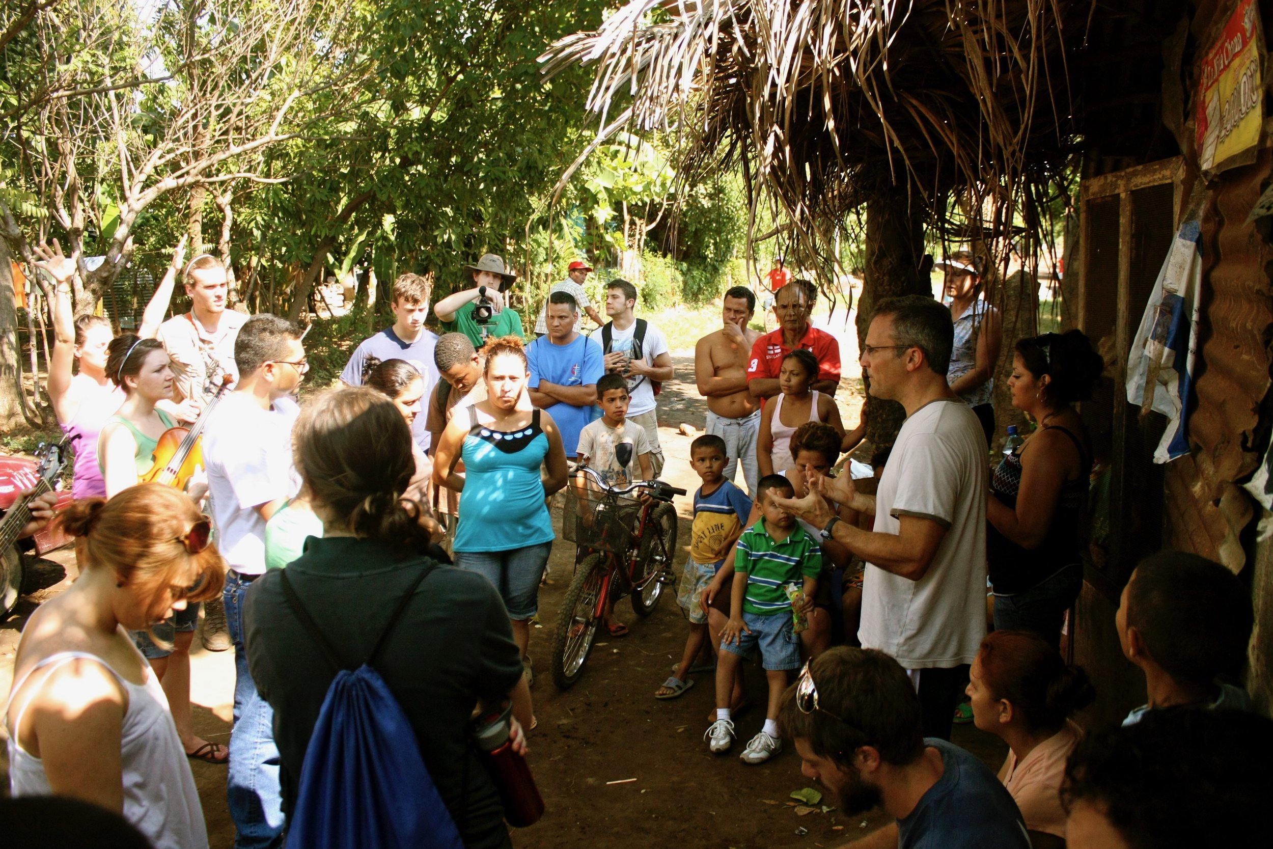 A man giving a speech or presentation outdoors to a group of diverse children and adults gathered around him in a shaded, rustic outdoor setting with trees and simple structures.