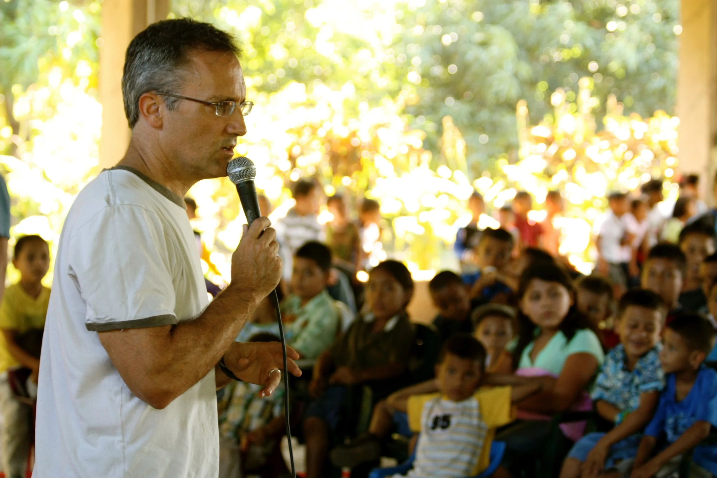 A man with glasses speaking into a microphone in front of a group of children indoors, with sunlight and trees visible outside.