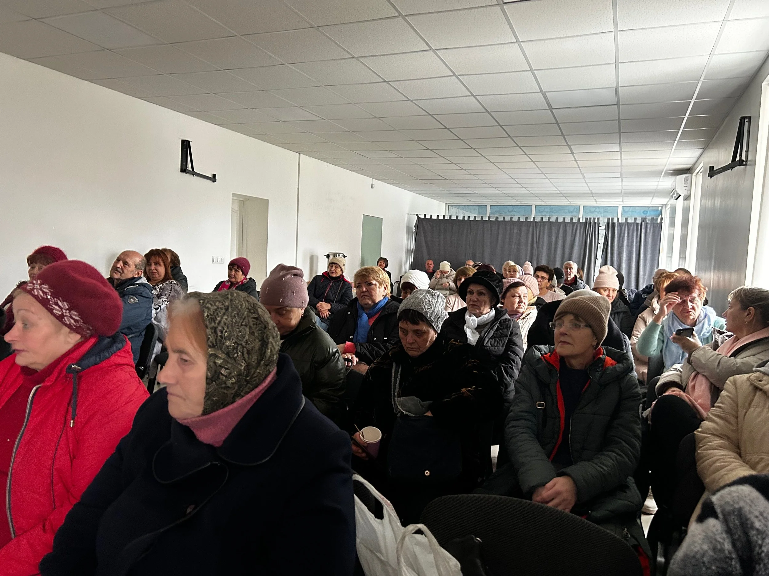 A group of elderly people sitting in chairs in a room, attentively listening to a presentation or speech. Many are wearing winter clothing and hats, with some holding cups or using phones.