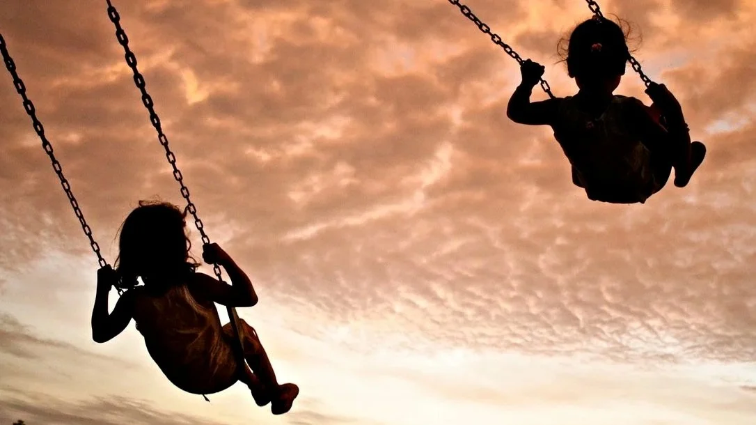 Silhouettes of two children on swings against an orange and pink sunset sky.