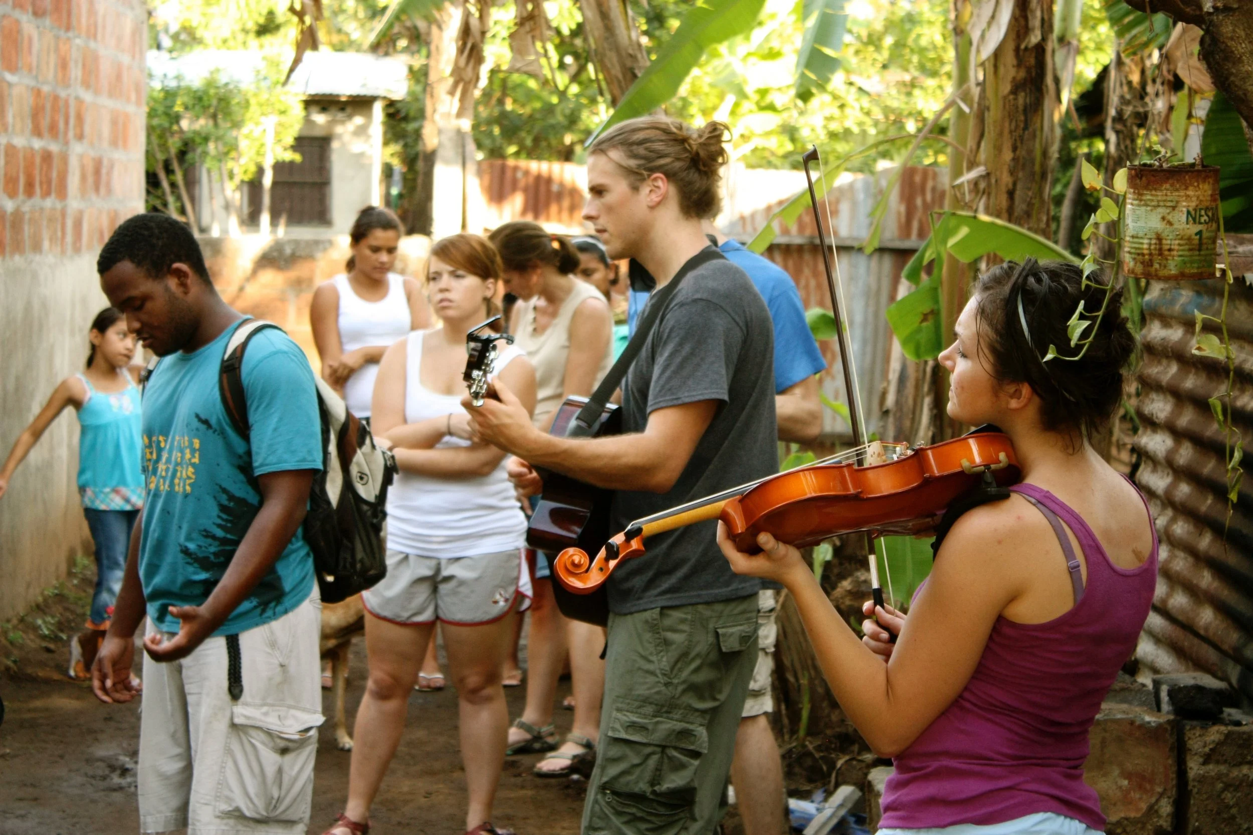 Street musicians performing for a small crowd in a backyard with a brick wall and green plants.