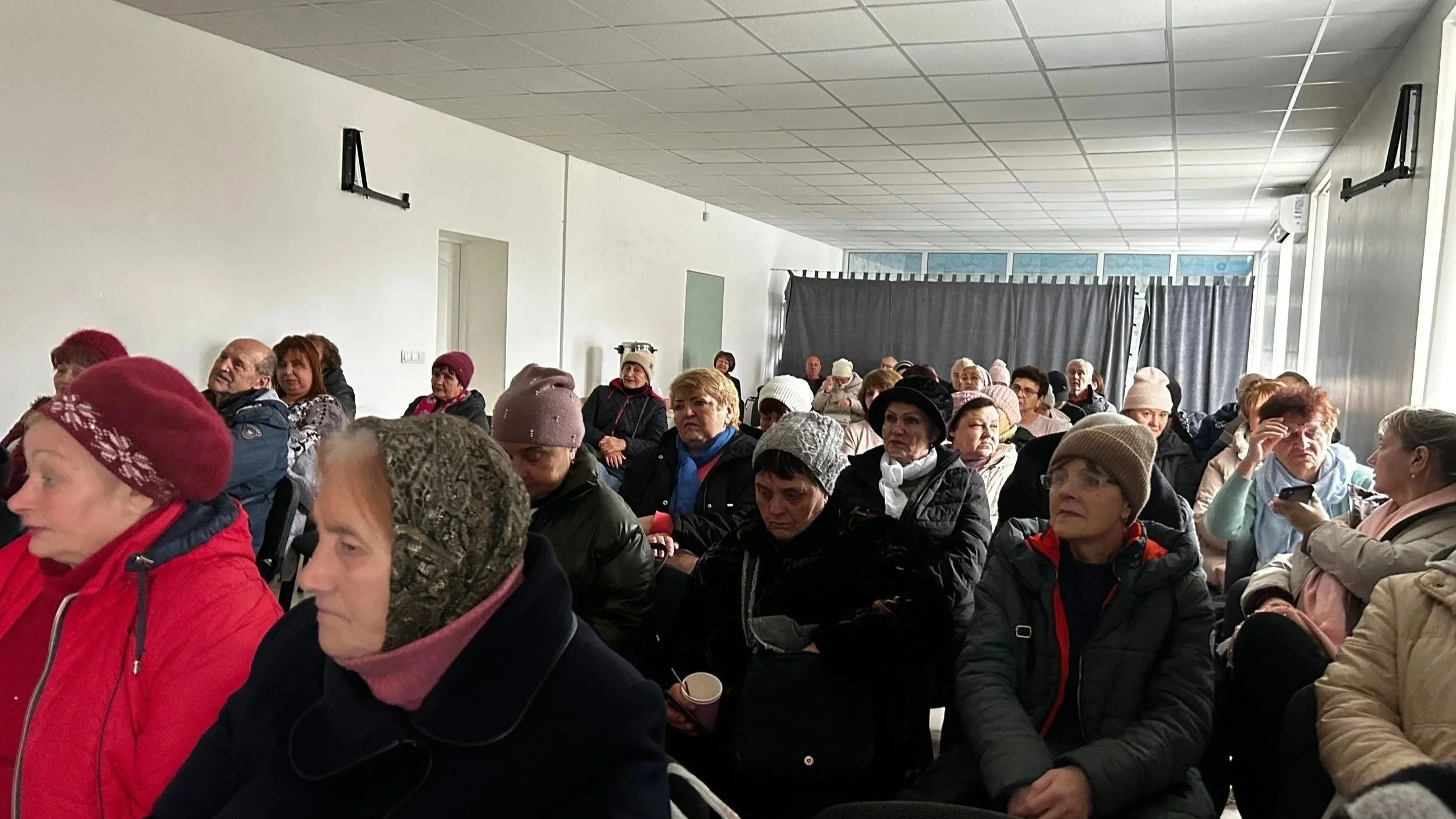 A group of elderly women and some men seated in a room with white walls and ceiling, many wearing winter hats and jackets, attending a gathering or presentation.