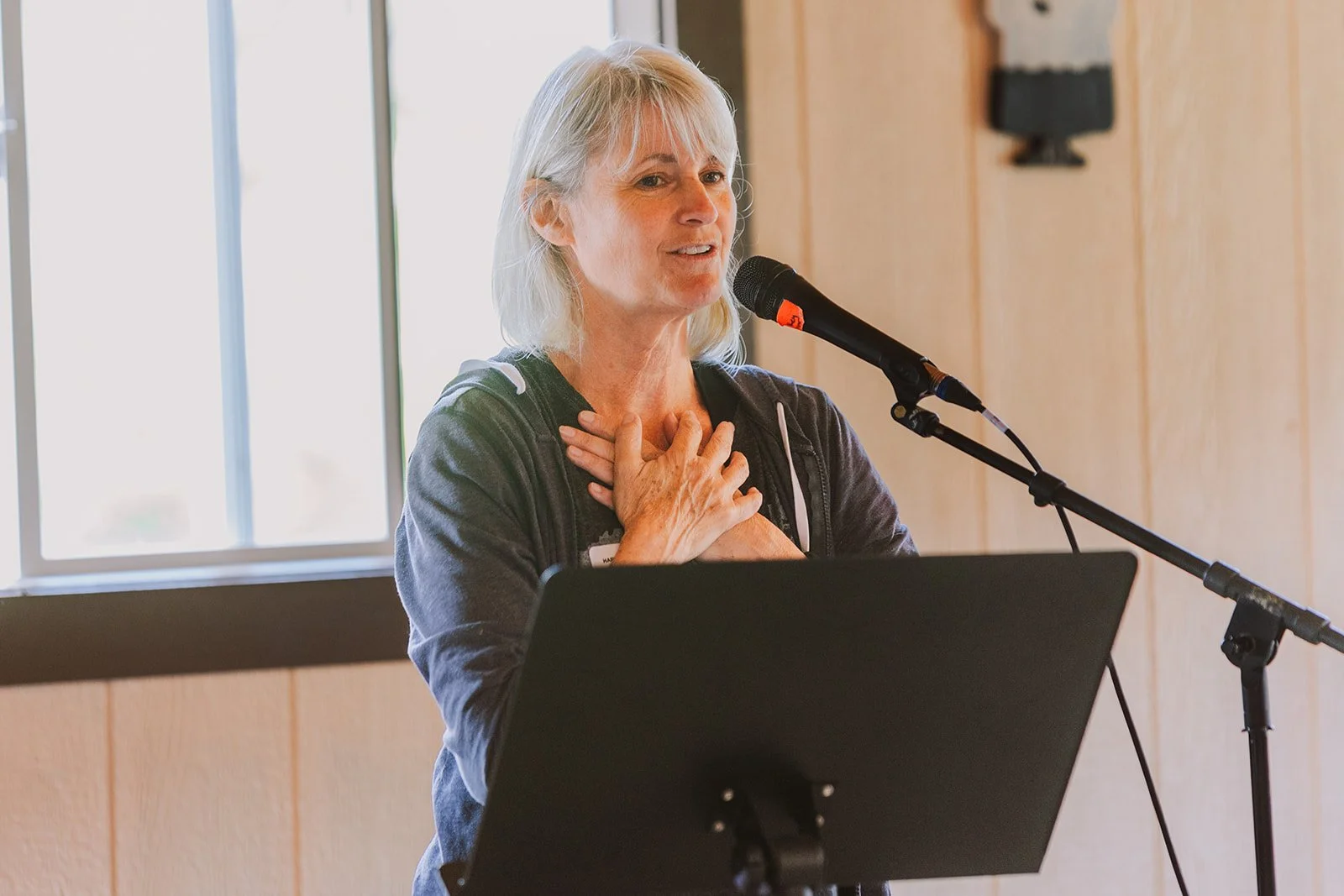 A woman with gray hair speaking into a microphone while standing behind a black music stand, with her hand on her chest, during a presentation or speech in a room with wood-paneled walls and a window.