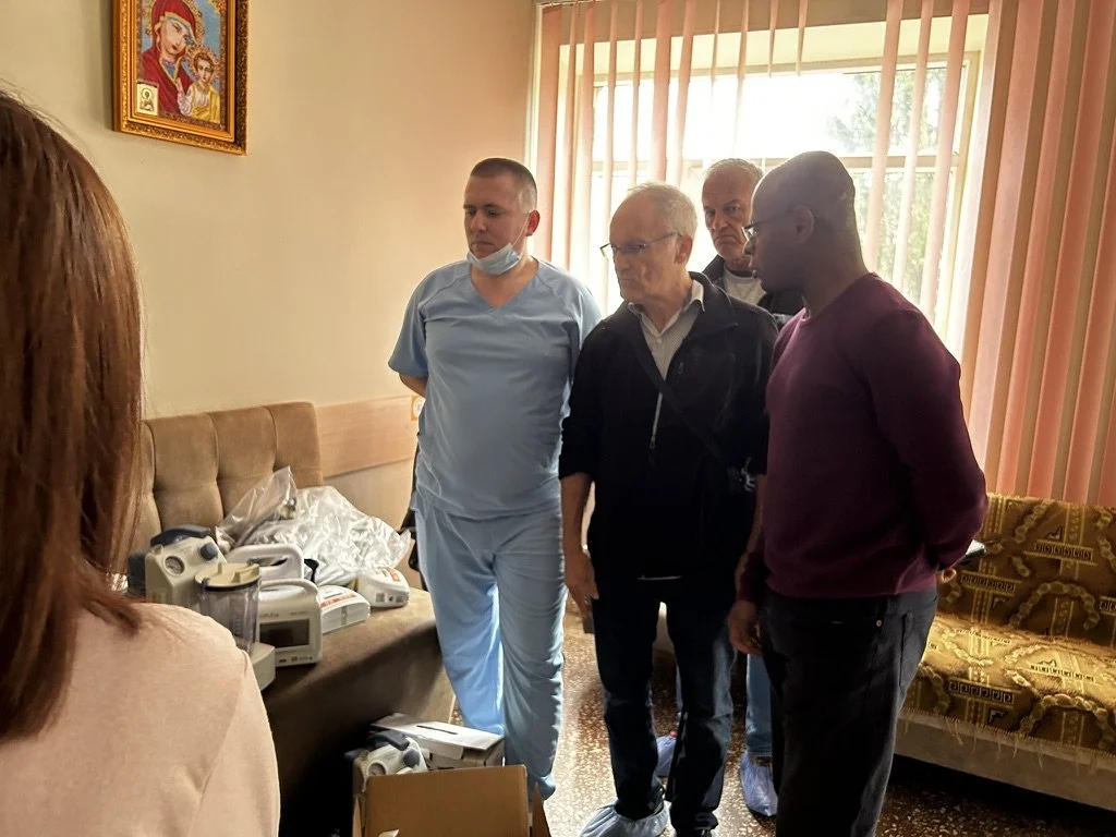 Group of five people standing in a room with medical equipment on a couch, looking at each other, with the woman in the foreground partially visible, a religious icon on the wall, and a window with pink vertical blinds.