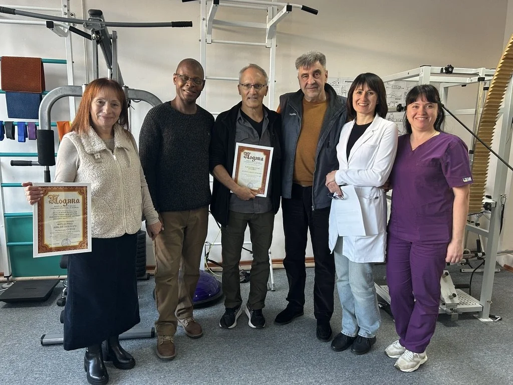 A group of six healthcare professionals standing together in a gym or therapy center, holding framed certificates and smiling at the camera.
