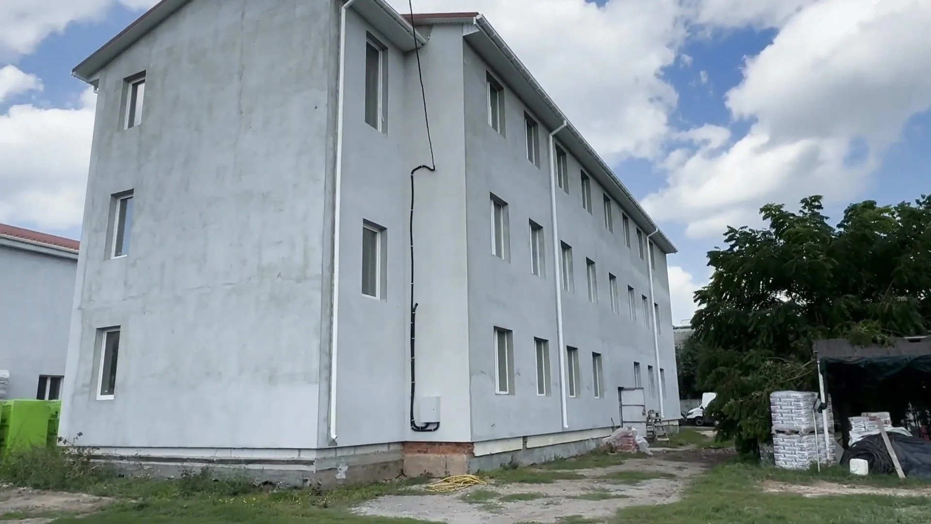 The side of a four-story building under construction with gray walls, multiple windows, and electrical wiring visible, surrounded by construction materials and equipment.