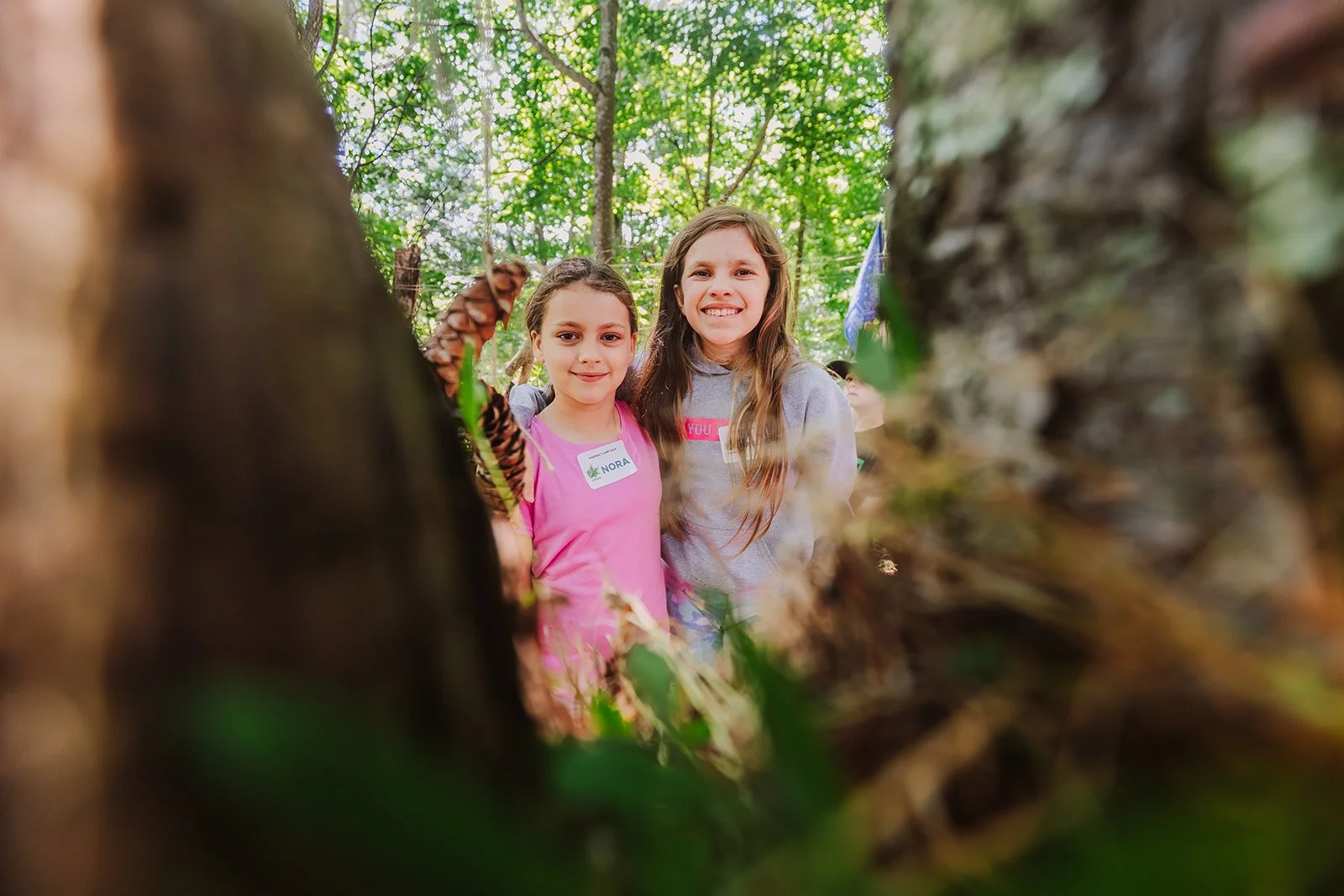 Two young girls standing in a wooded forest, smiling at the camera, seen through a hole in a fallen tree branch.