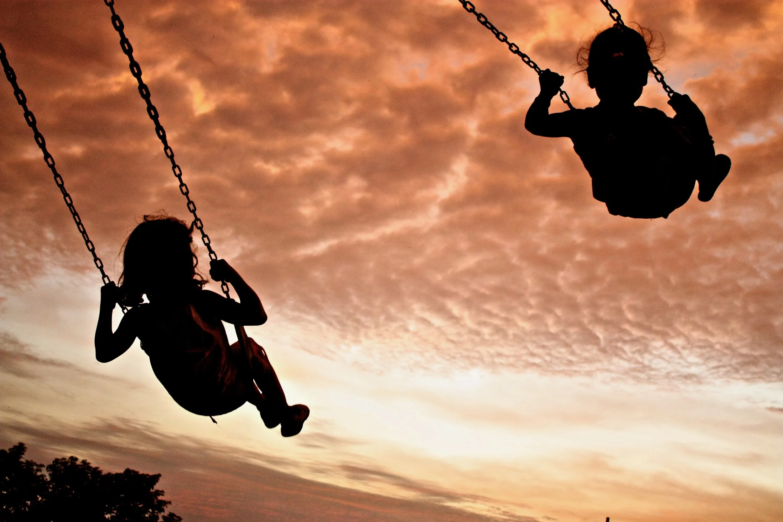 Silhouettes of two children on swings against a sunset sky with orange clouds.
