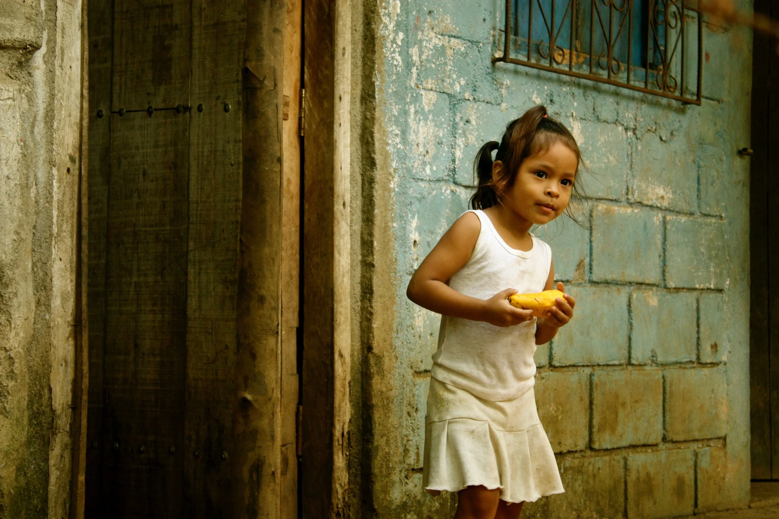 A young girl holding a yellow object, standing in front of a weathered blue wall with a window and a wooden door, in a rustic setting.