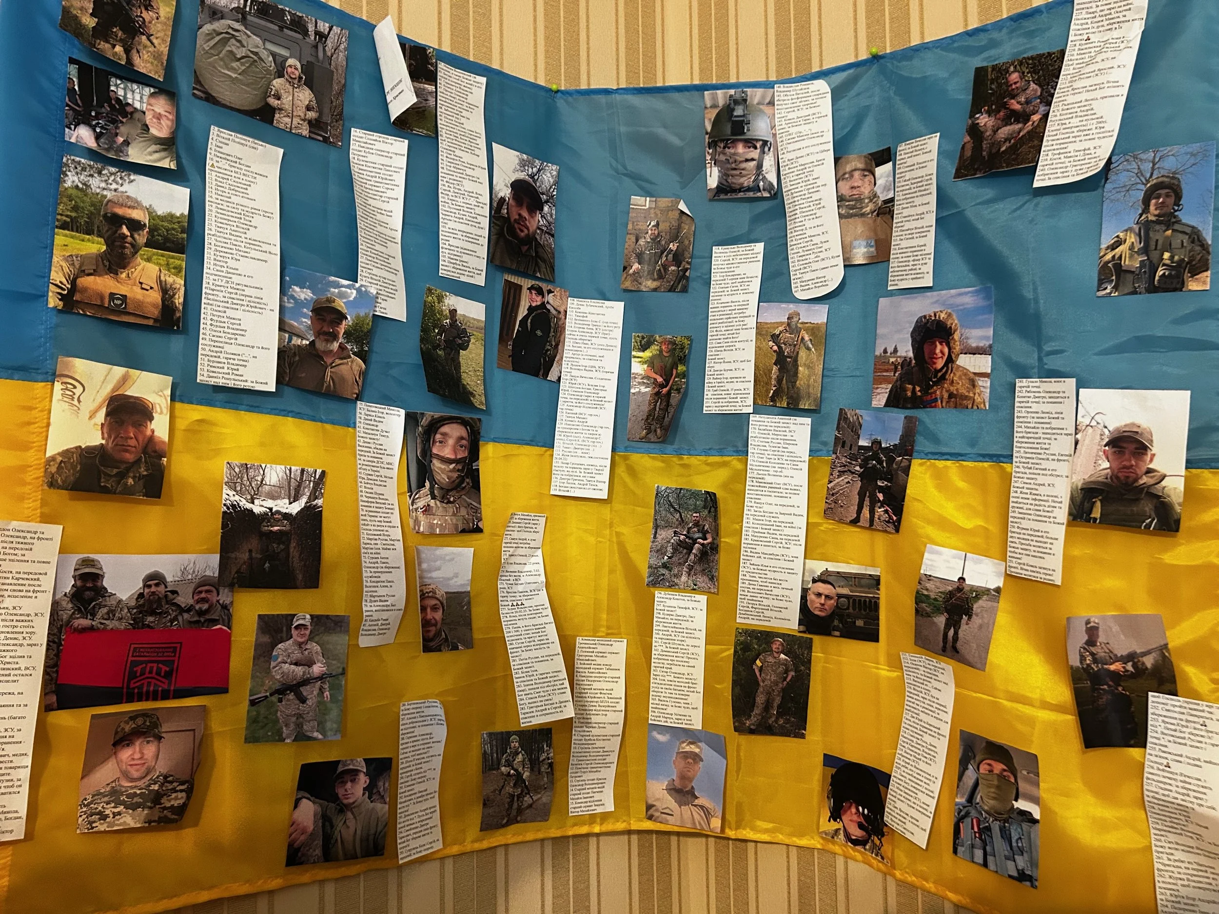 Display board with numerous photographs of soldiers and accompanying written descriptions, featuring the Ukrainian flag in the background.