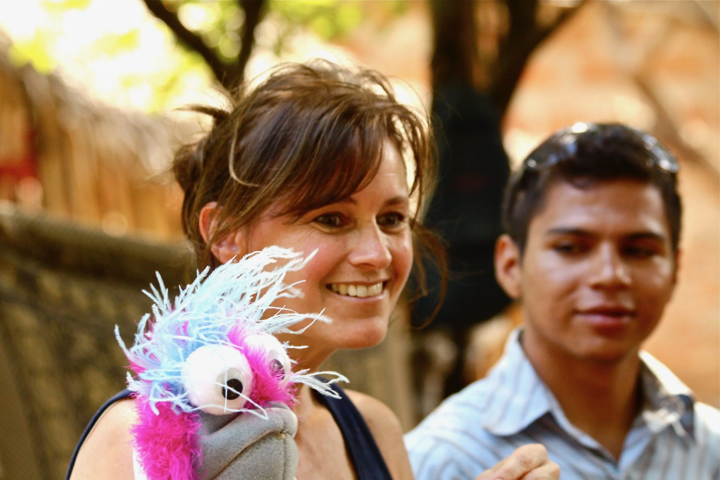 Smiling woman holding a colorful plush puppet with large eyes, with a young man in the background, outdoors with trees.