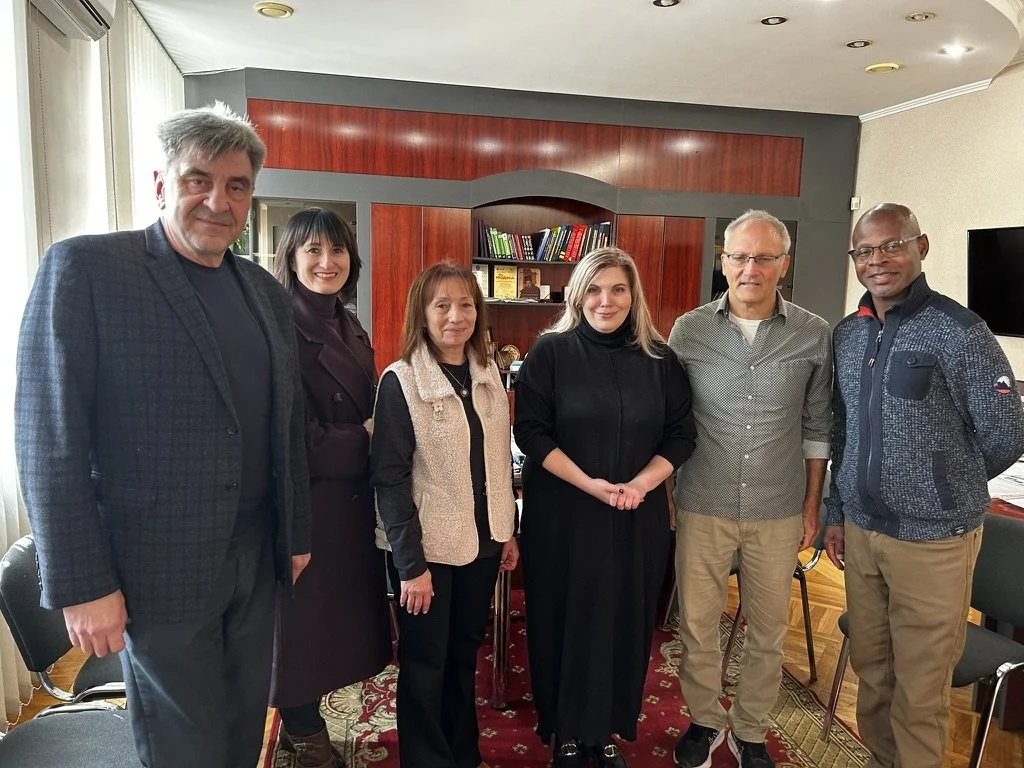 Six people standing together indoors in a room with bookshelves and a television, posing for a photo.