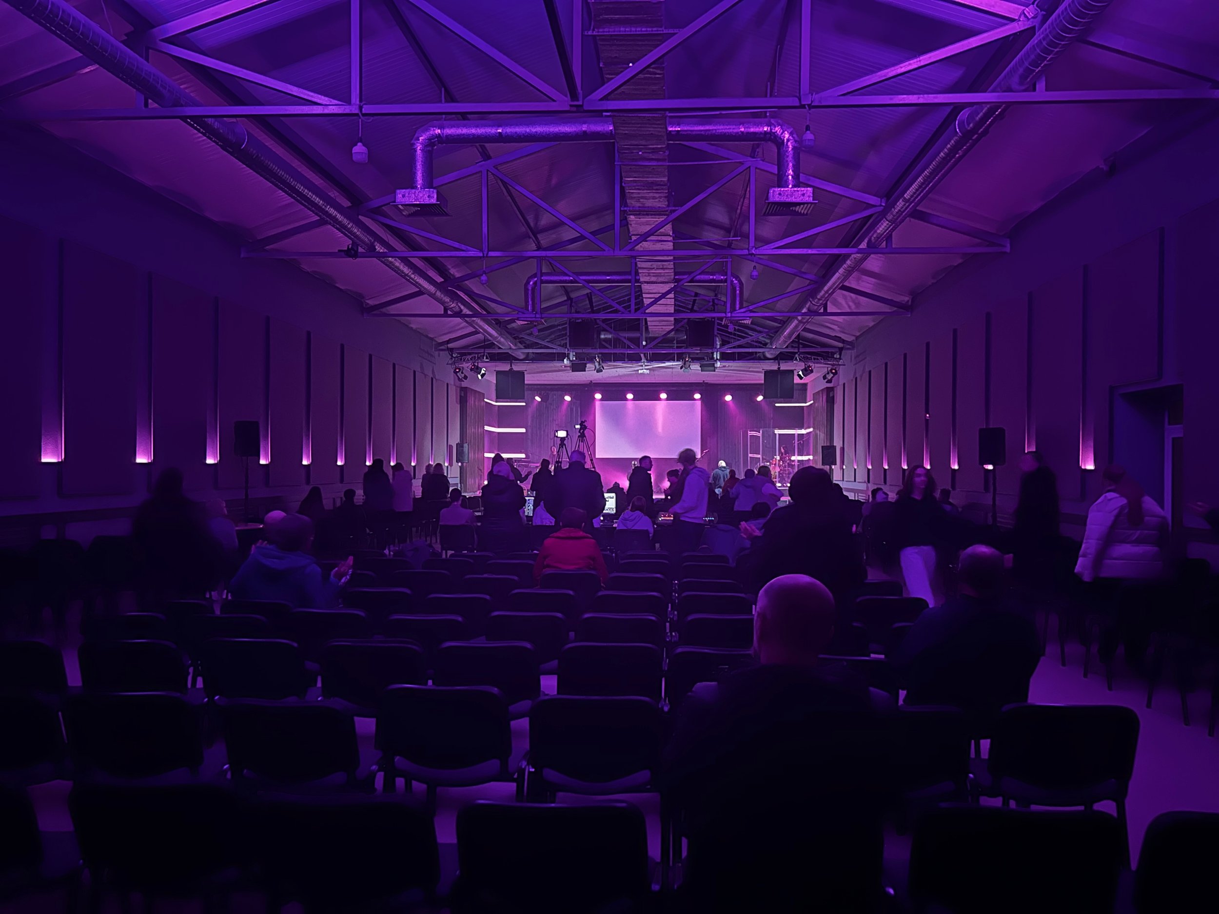 An indoor event space with purple lighting, a stage at the front with equipment, and people seated and standing, preparing for a performance or presentation.