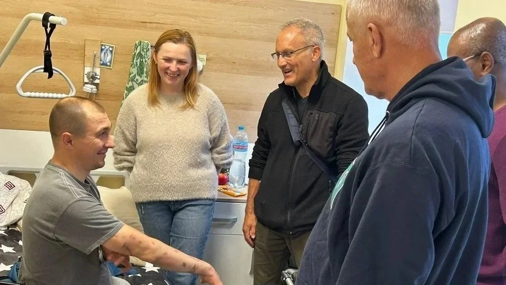 A group of five people in a hospital room, with one person in a hospital bed. The person in the bed is smiling and talking to the others, who are standing and engaged in conversation. One person is wearing a beige sweater, and others are in dark or colorful hoodies.