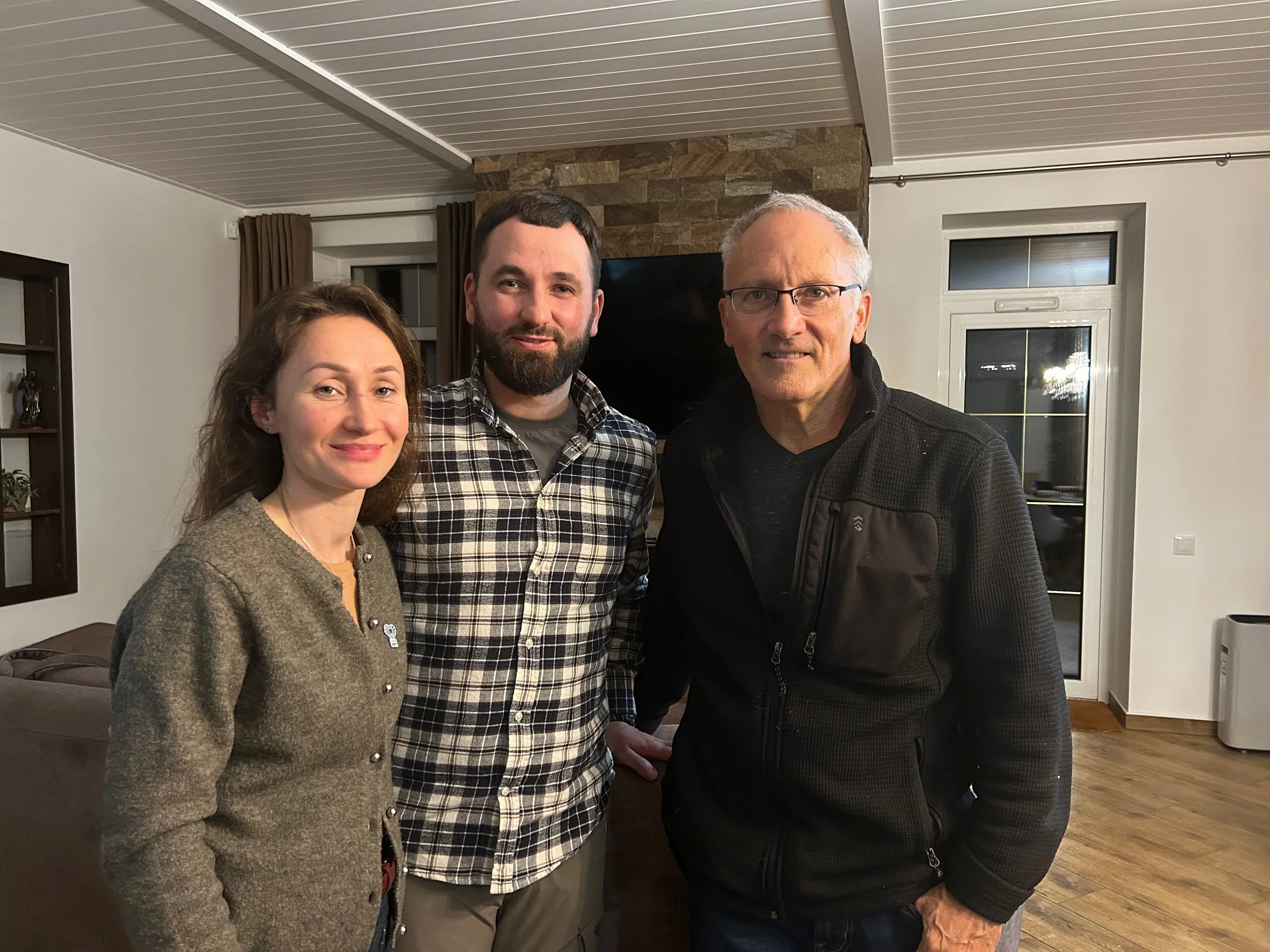Three people standing indoors in front of a television, smiling at the camera.