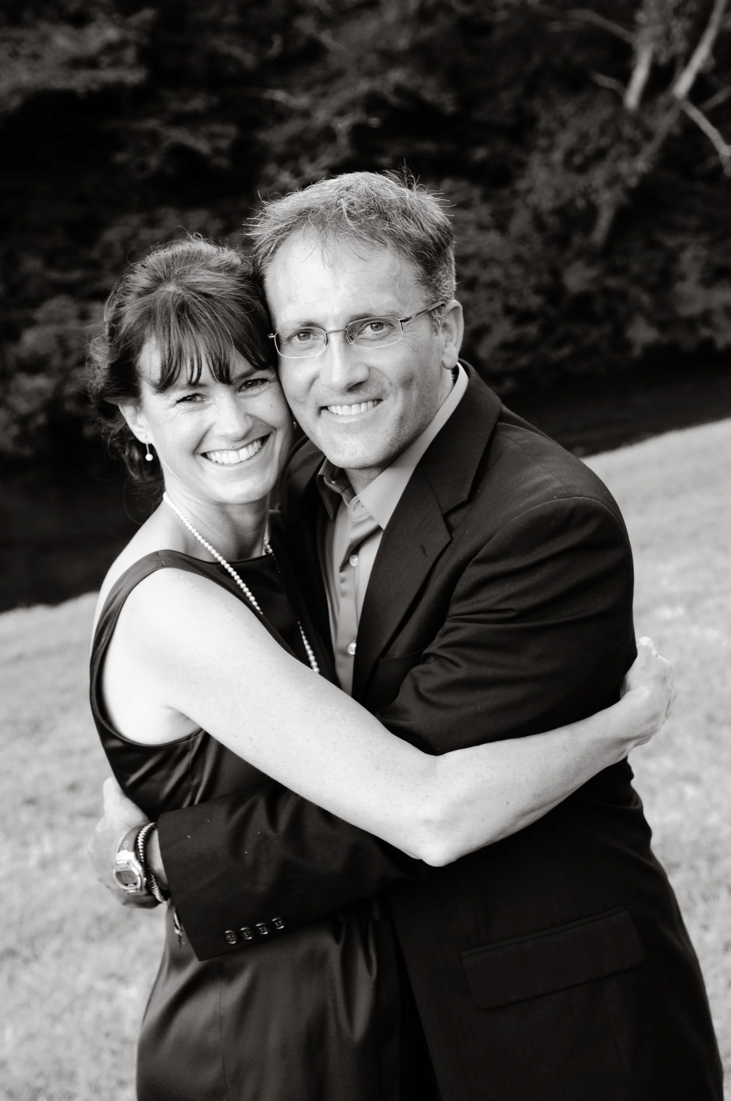 A smiling man and woman hugging outdoors near a river, in black and white.