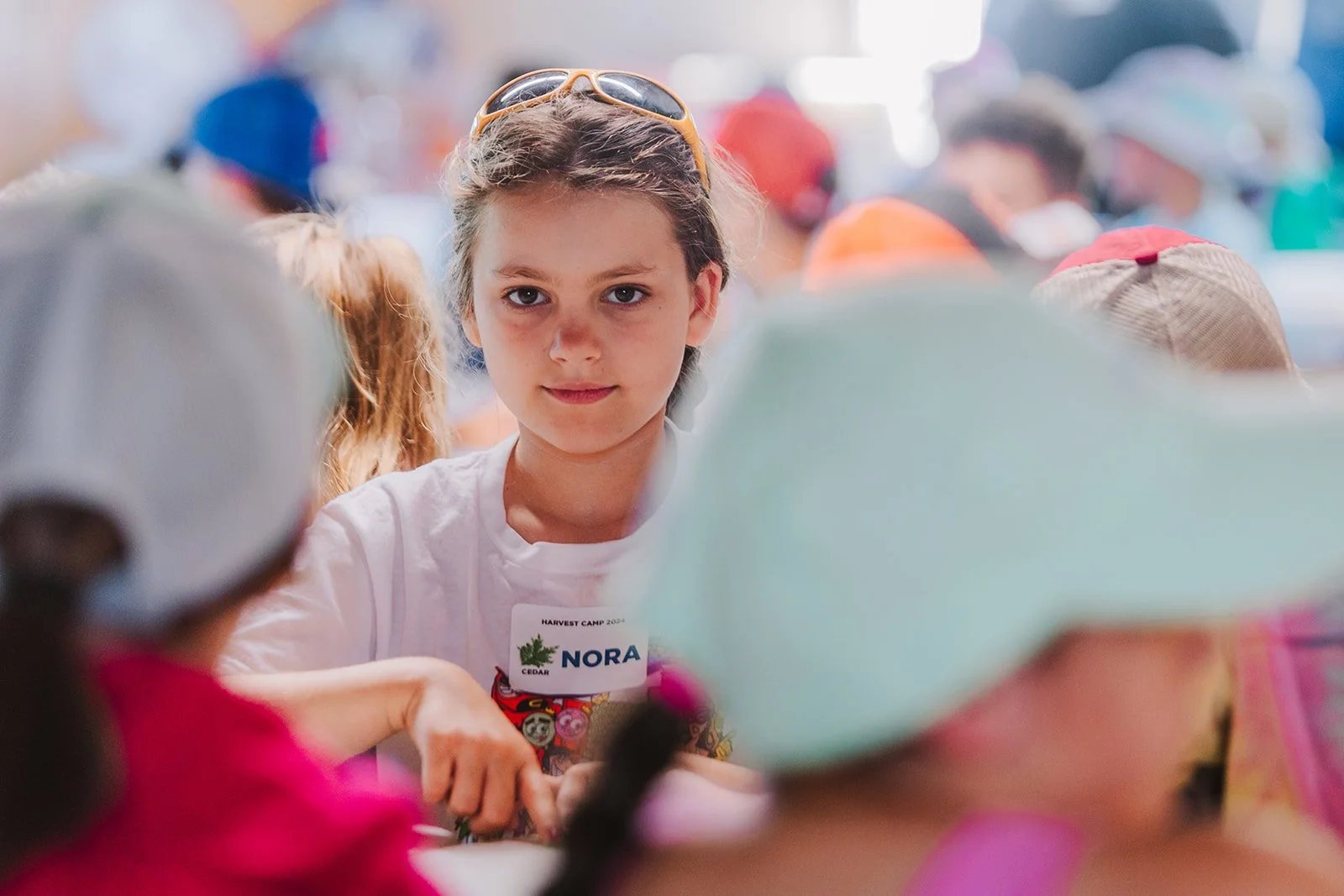 A young girl with brown hair and sunglasses on her head, looking at the camera, surrounded by other children at a camp or event.
