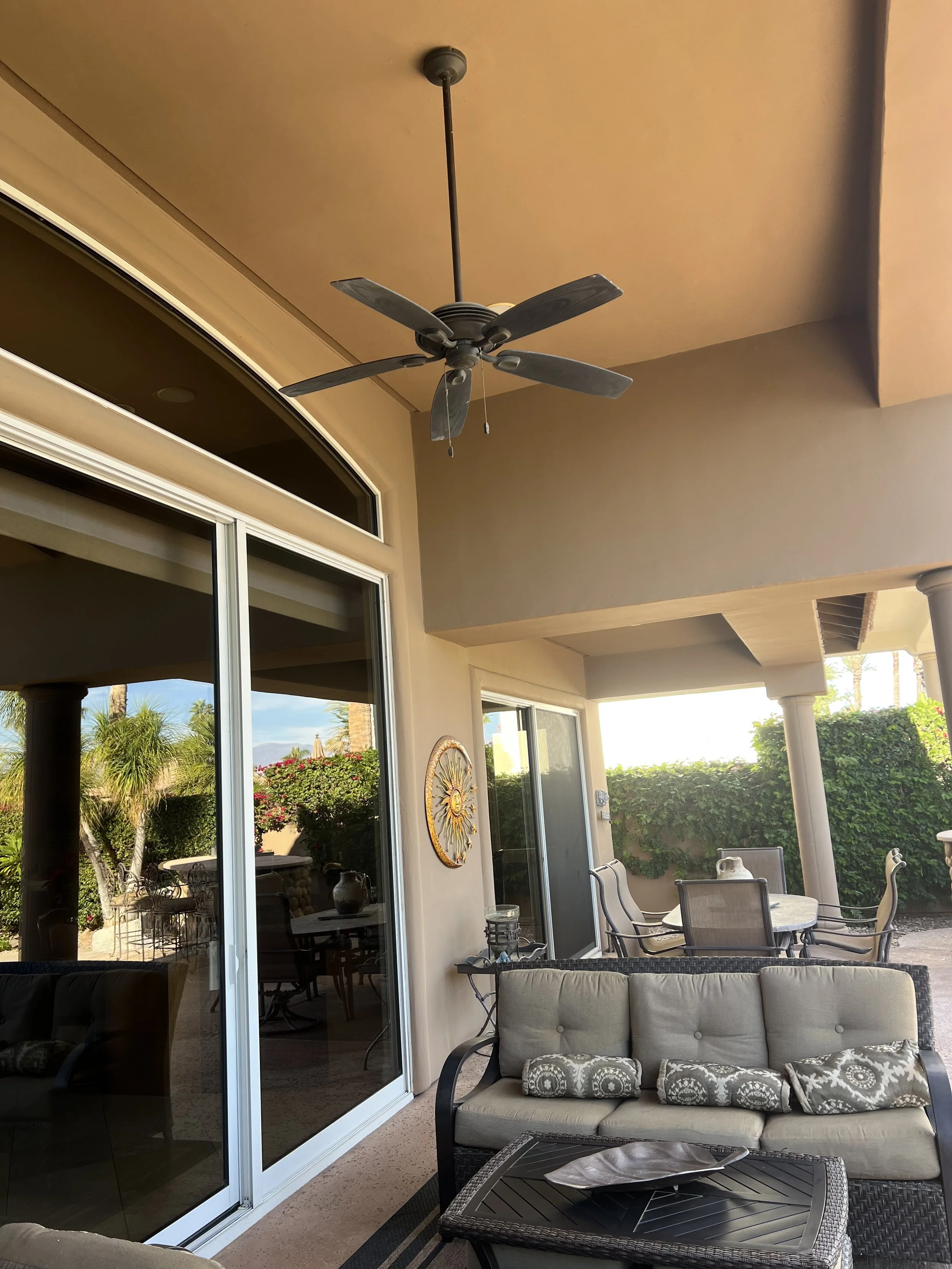 Covered outdoor patio with ceiling fan, sliding glass doors, beige cushioned seating, and garden view with trees and bushes.