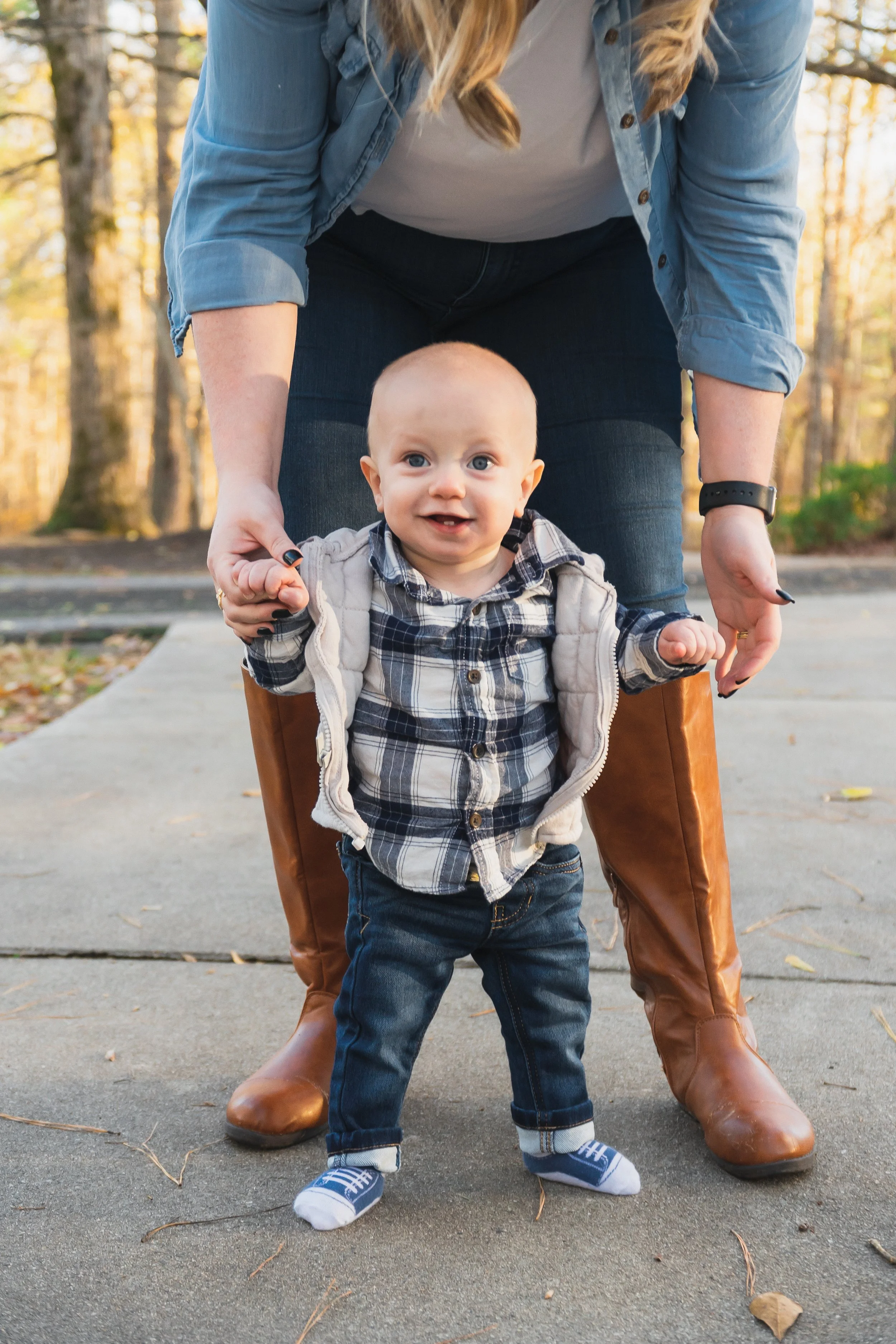 A smiling baby with blue eyes standing outdoors on a sidewalk, holding hands with a woman bending down behind him, surrounded by trees with autumn leaves.