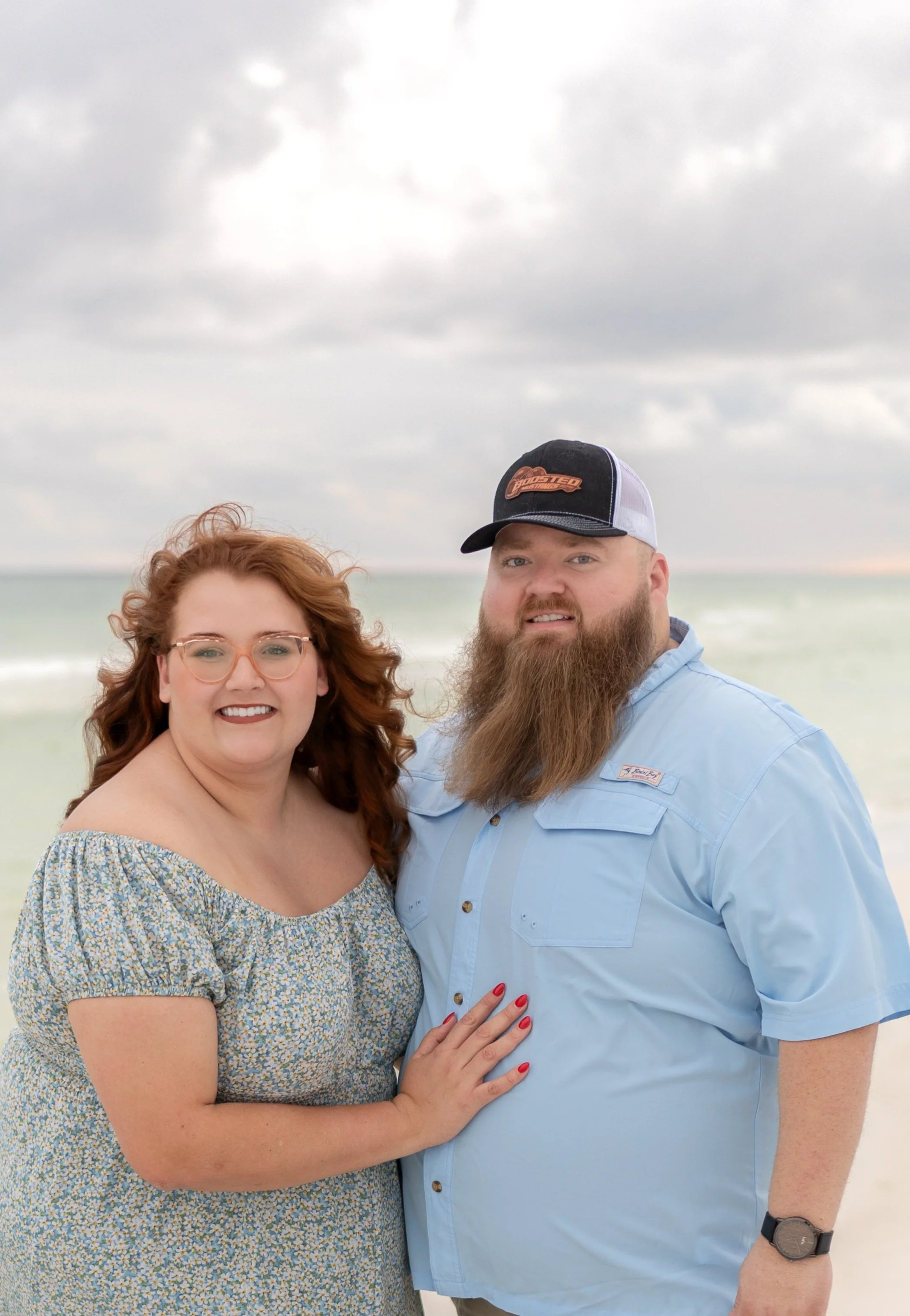 A smiling woman and man posing together on a beach with cloudy sky and ocean in the background.