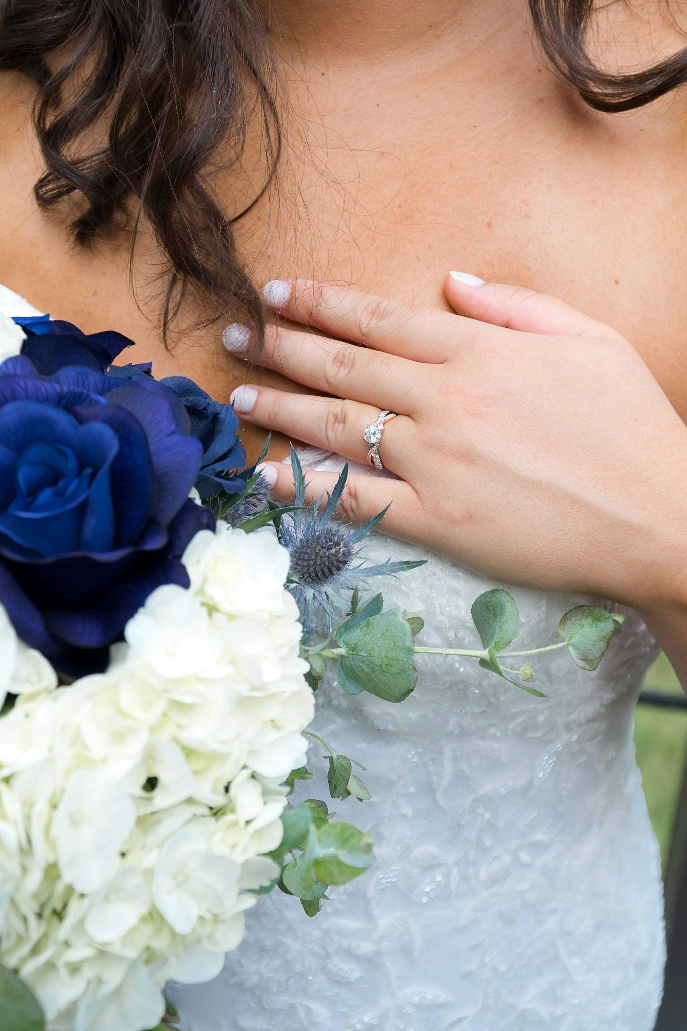 Close-up of a bride touching her neck, showing a diamond engagement ring, with a bouquet of blue and white flowers in the foreground.