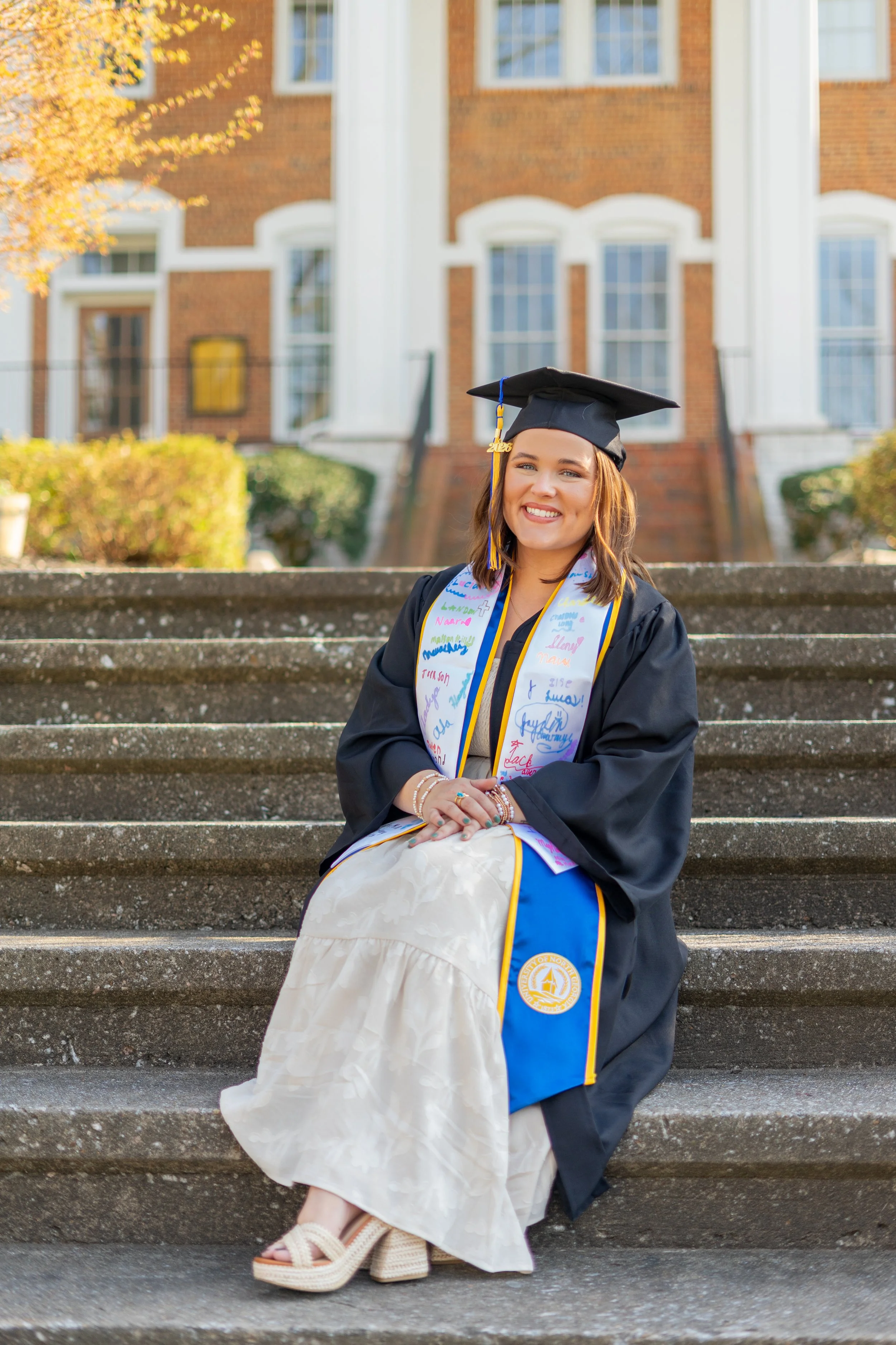 A young woman in a black graduation gown and cap sits on outdoor steps, smiling, with a white stole with handwritten signatures. She wears sandals and has a diploma. A brick building with large windows is in the background, along with a tree with yel