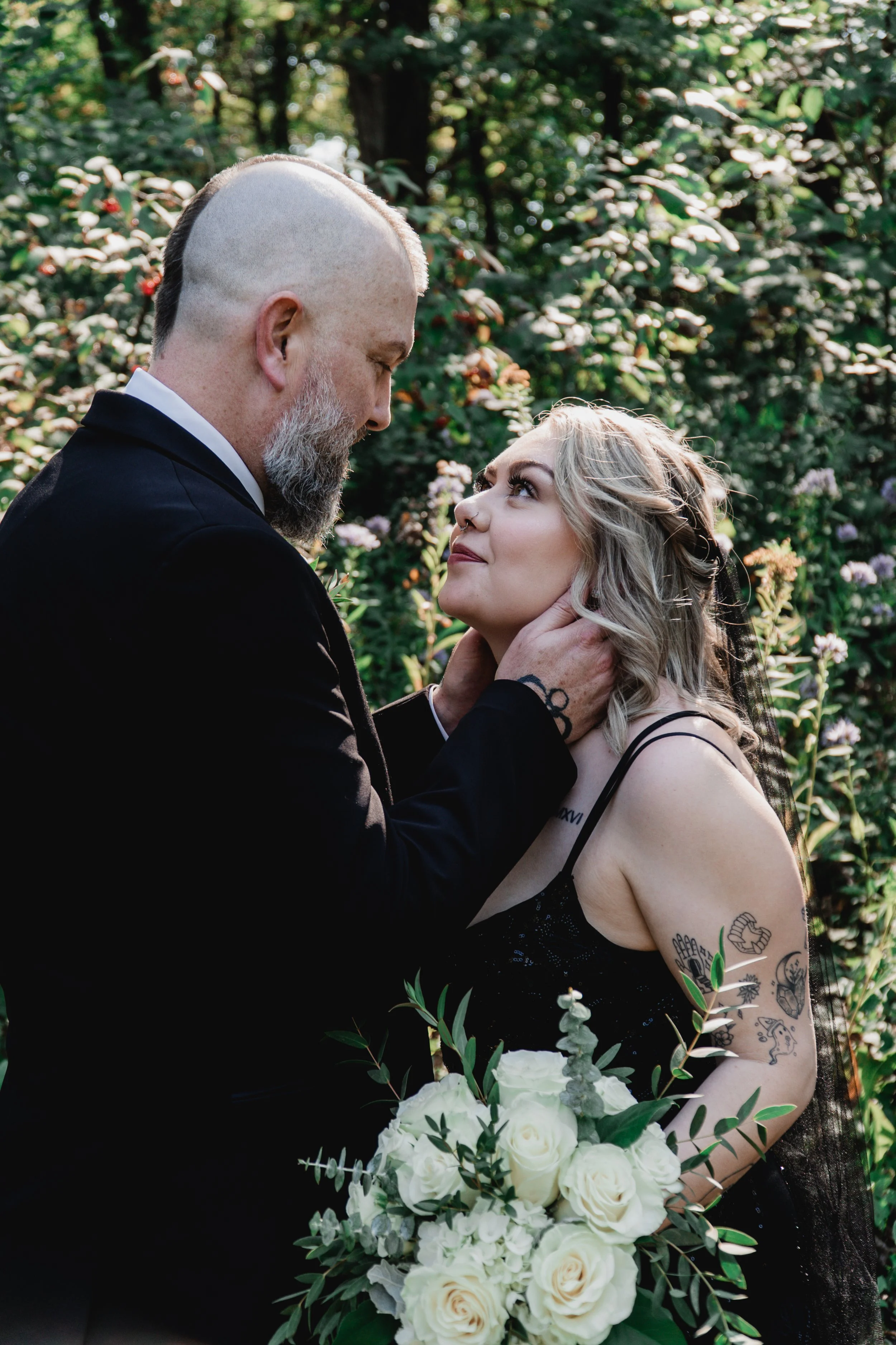 A man and a woman in an outdoor setting, face to face. The man has a beard, shaved sides, and is wearing a black jacket and a white shirt. The woman has blonde curly hair, tattoos on her arms, and is holding a bouquet of white roses and greenery. The
