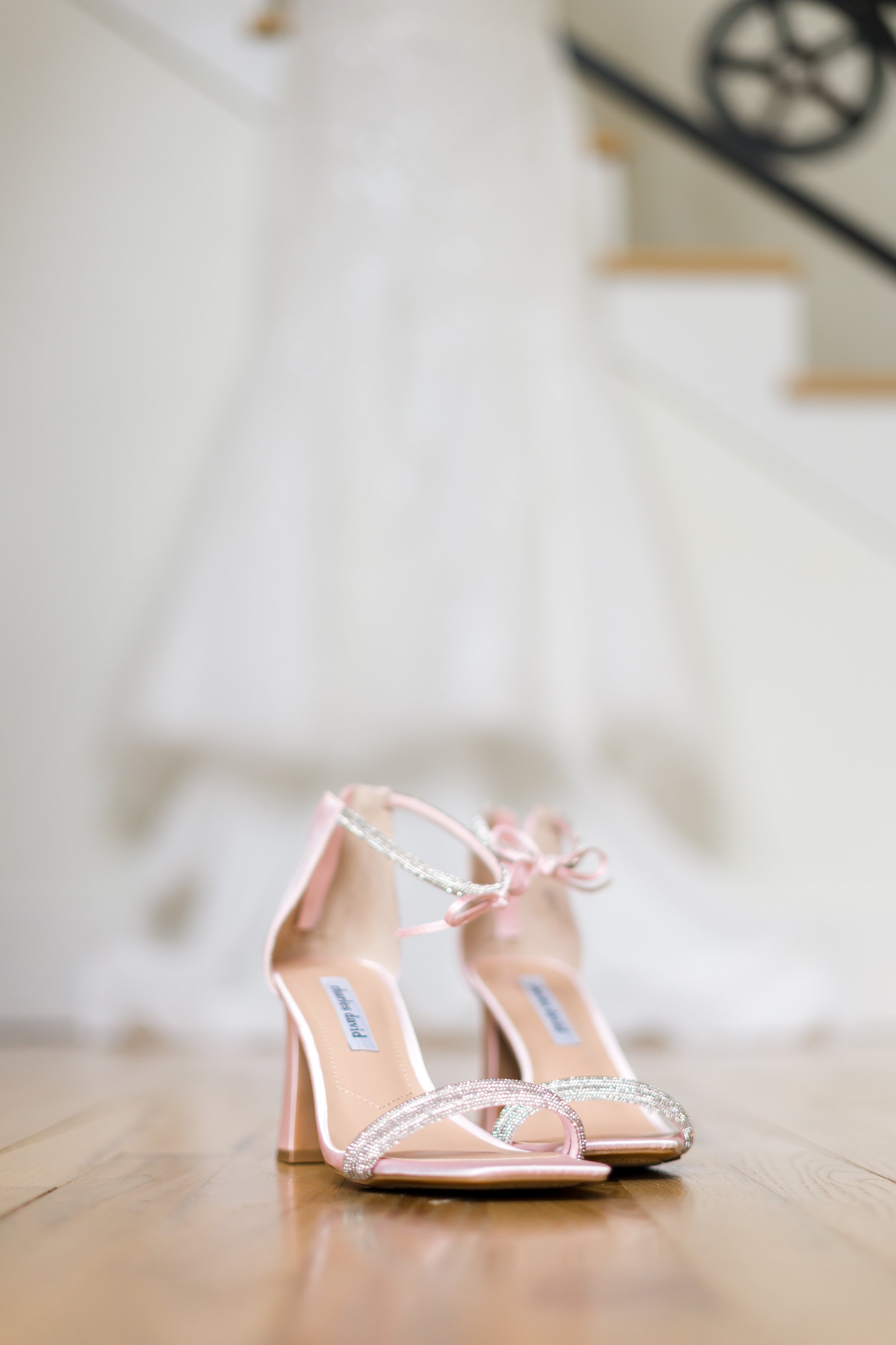 Pink high-heeled sandals with rhinestones on a wooden floor with a white wedding dress hanging in the background.