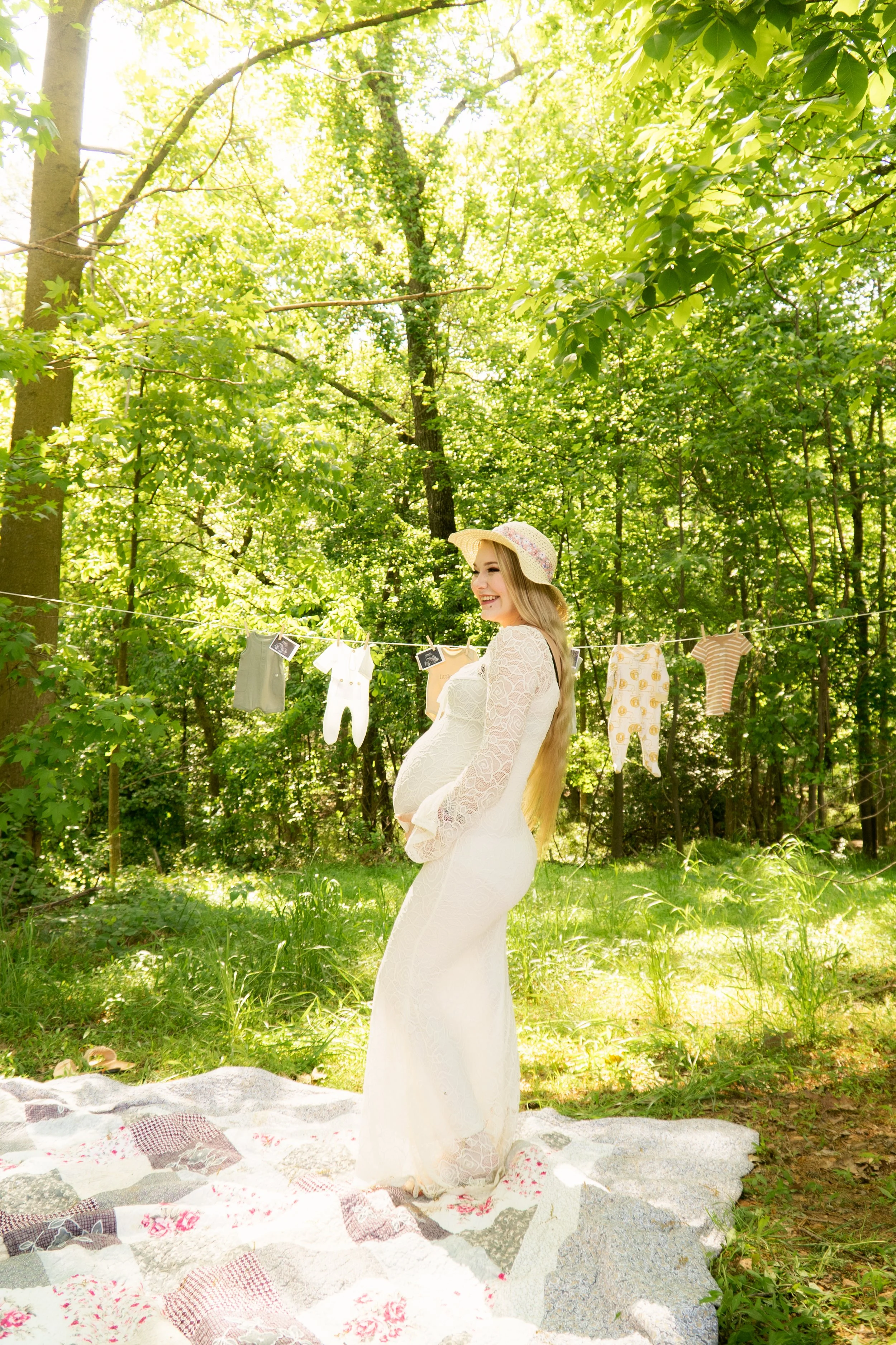 Pregnant woman in a white lace dress holding her belly outdoors in a forest, with laundry hanging on a clothesline behind her, bright sunlight filtering through green leaves.