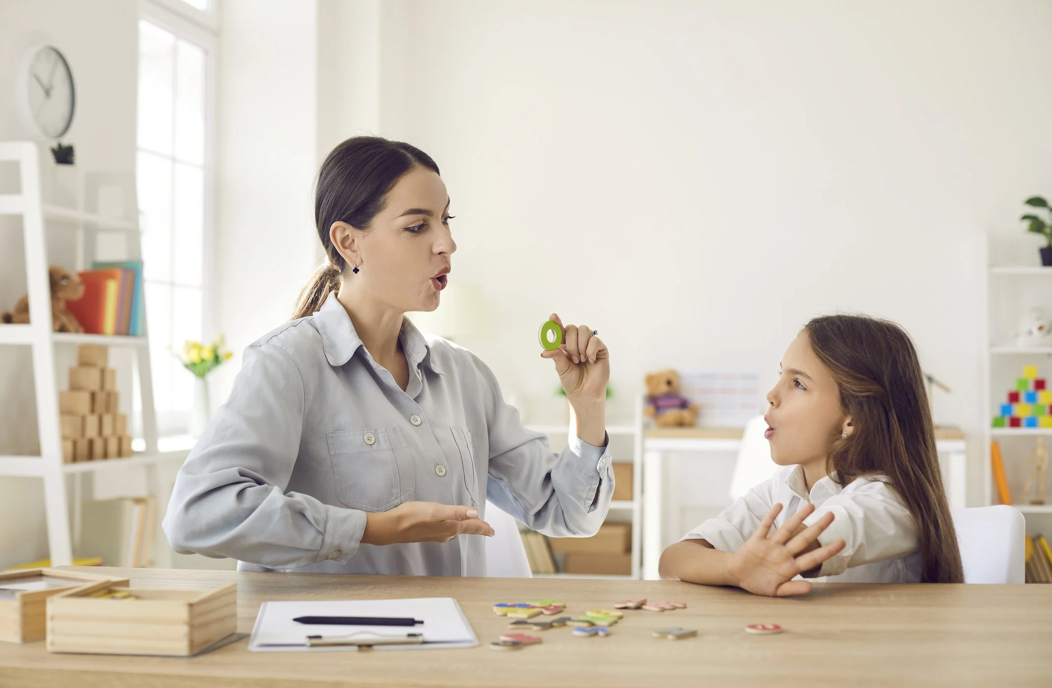 A woman and a girl are engaged in a learning activity in a bright classroom. The woman is holding a green puzzle piece, and the girl is gesturing with her hands, with puzzle pieces on the table in front of them.