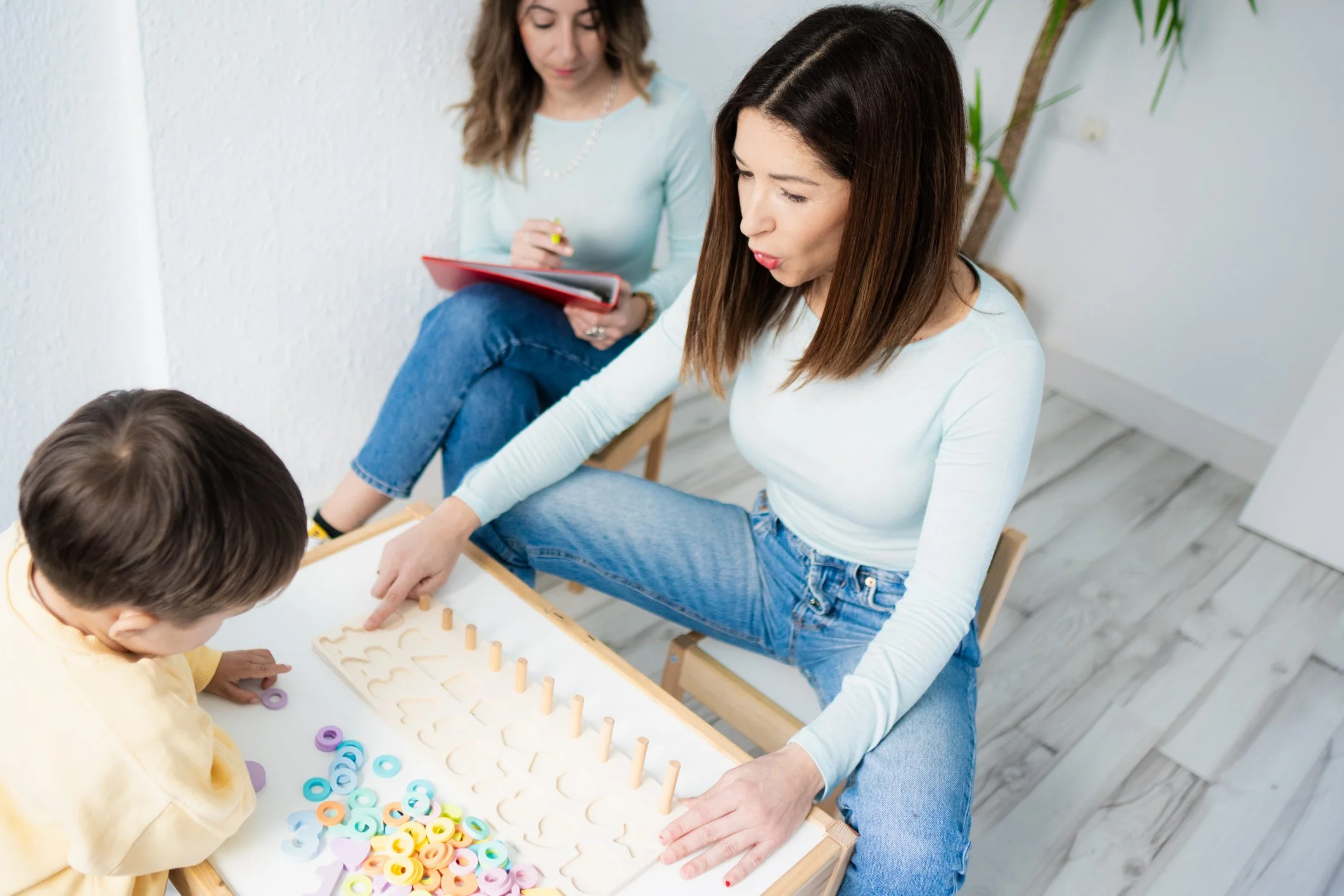 Two women and a young girl playing a tabletop game with colorful shaped pieces, one woman is sitting and pointing at the game while the other woman and girl watch.